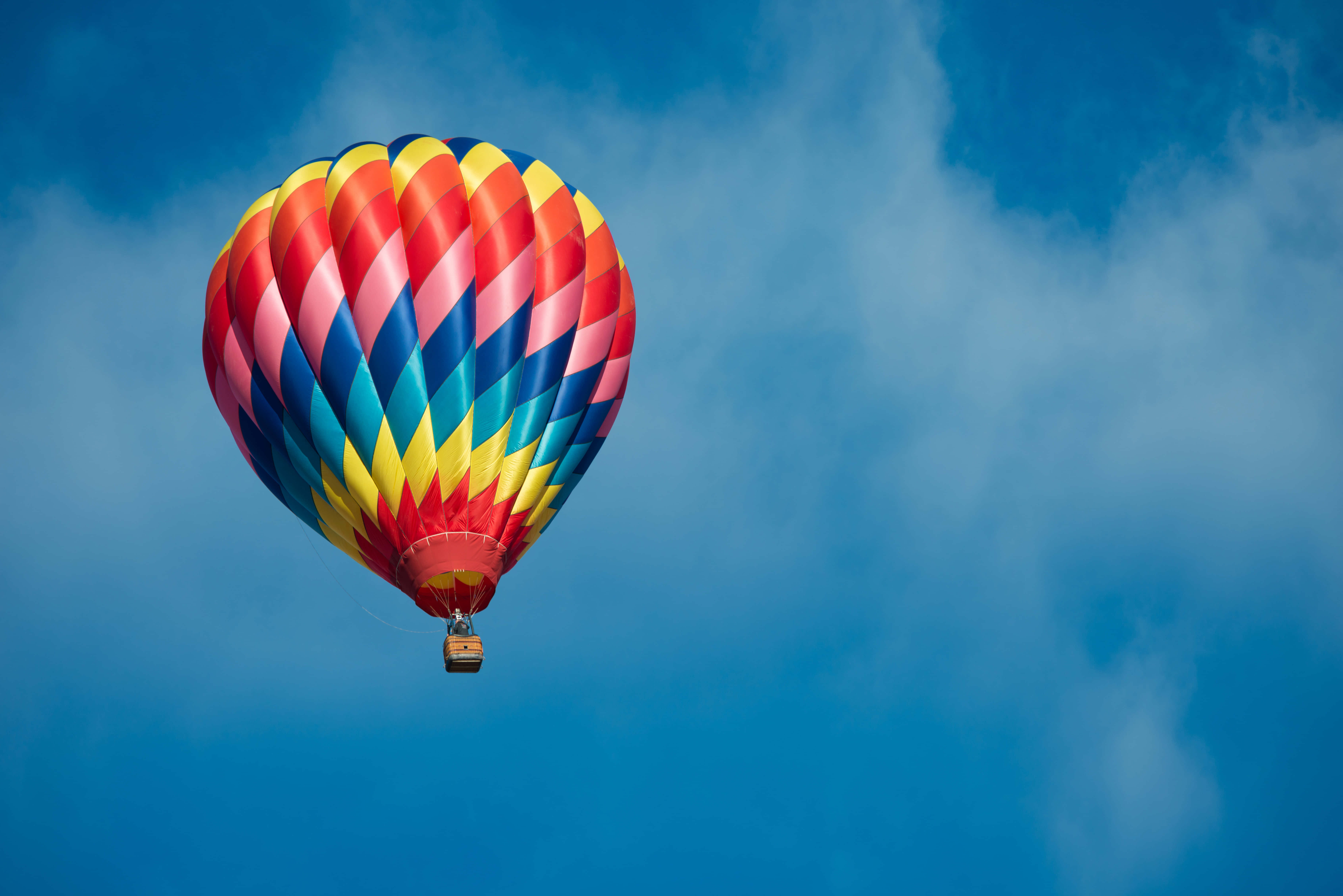 Hot Air Balloon 桌布 壁紙 — 一個色彩鮮豔的熱氣球，帶有紅色、藍色和黃色條紋，在晴朗的藍天中伴隨著輕柔的雲朵靜靜漂浮。