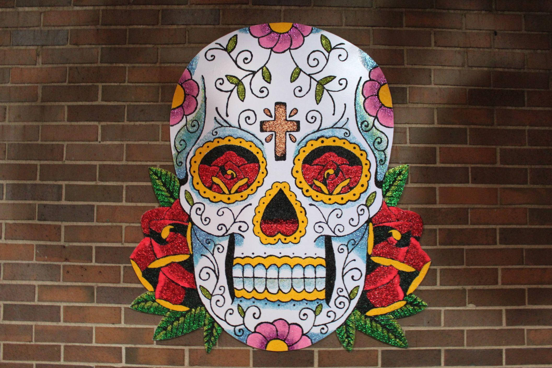 Dia De Los Muertos 桌布 壁紙 — 一個色彩鮮豔的糖骷髏，飾有玫瑰和漩渦圖案，掛在紅磚牆上。