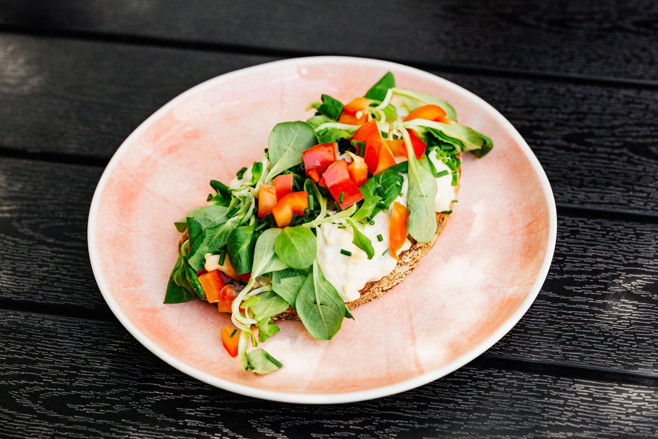 Healthy Food 桌布 壁紙 — 一個色彩鮮豔的開放式三明治，上面有奶油白色塗抹醬、新鮮蔬菜和紅椒丁，放在柔軟的粉紅色盤子上，置於深色木板上。