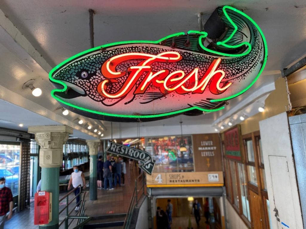 Pike Place Market 桌布 壁紙 — 一個色彩鮮豔的魚形霓虹燈牌，上面有草書字體的「Fresh」，在繁忙的室內市場走廊上方發光。