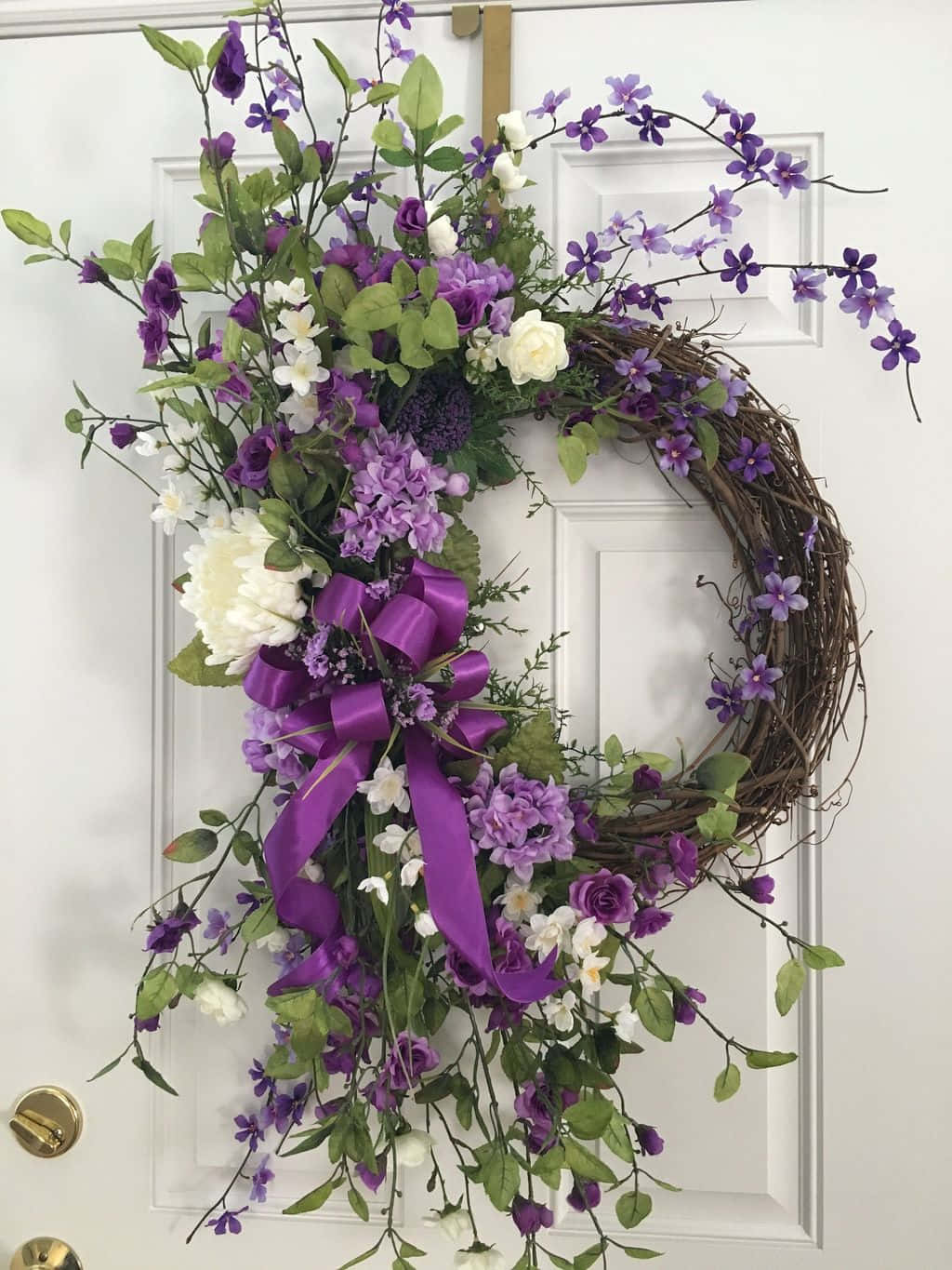 Spring Wreaths 桌布 壁紙 — 一個茂密的紫色和白色花環，配有綠色植物和一個大蝴蝶結，掛在白色門上。