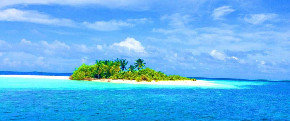 Maldives Island 桌布 壁紙 — 一個茂密的綠色熱帶島嶼，棕櫚樹矗立在白色沙洲上，周圍是充滿活力的碧綠海水，上方是明亮的藍天，點綴著幾朵白雲。