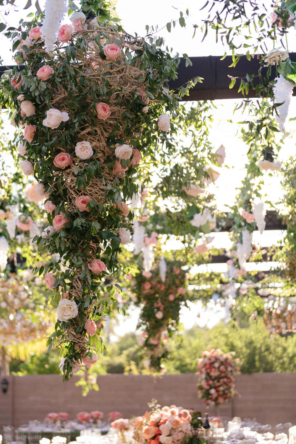Wedding Aesthetic 桌布 壁紙 — 一個茂盛的花拱門，掛滿粉紅色和白色的玫瑰和綠葉，懸掛在陽光明媚的戶外婚禮招待會上方。
