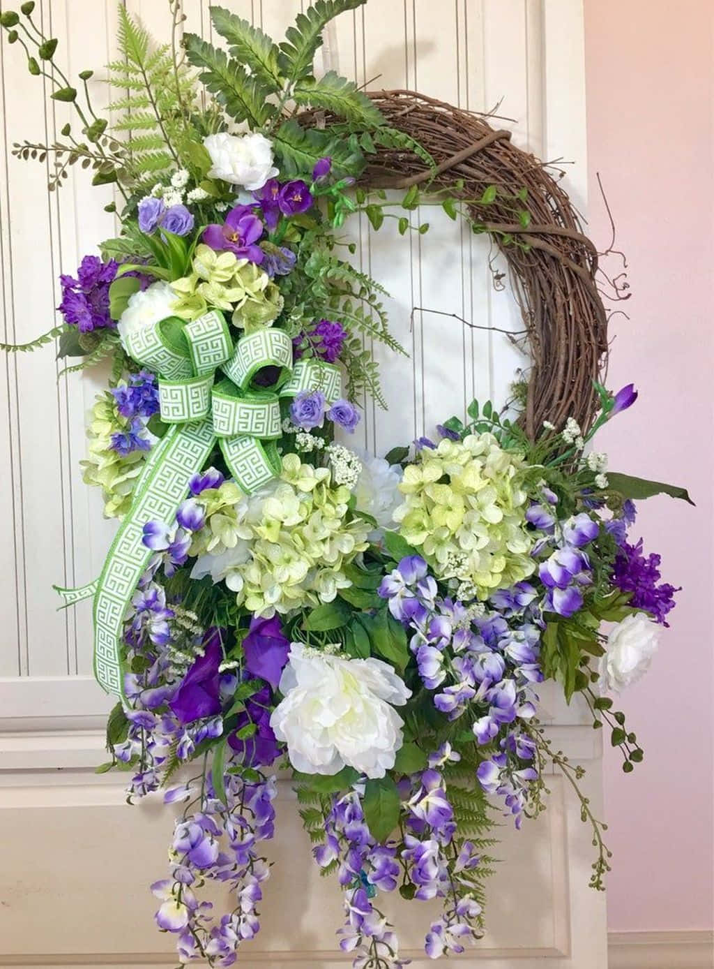 Spring Wreaths 桌布 壁紙 — 一個茂盛的花環，有紫色紫藤、綠色繡球花和白色玫瑰，掛在白色鑲板門上，背景是柔和的粉紅色牆壁。