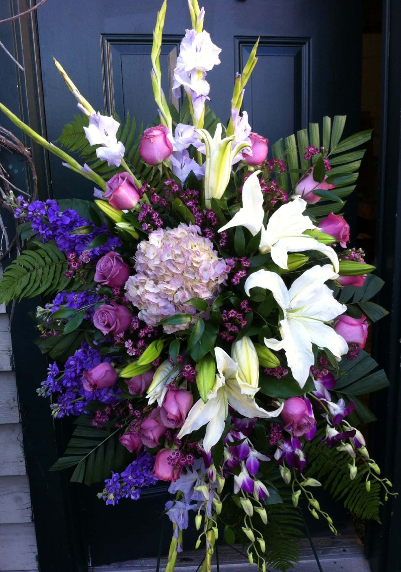 Funeral Flower Arrangements 桌布 壁紙 — 一個茂盛的花藝佈置，包含白色百合、粉紅玫瑰、紫色蘭花和繡球花，在深色門前優雅地垂墜。
