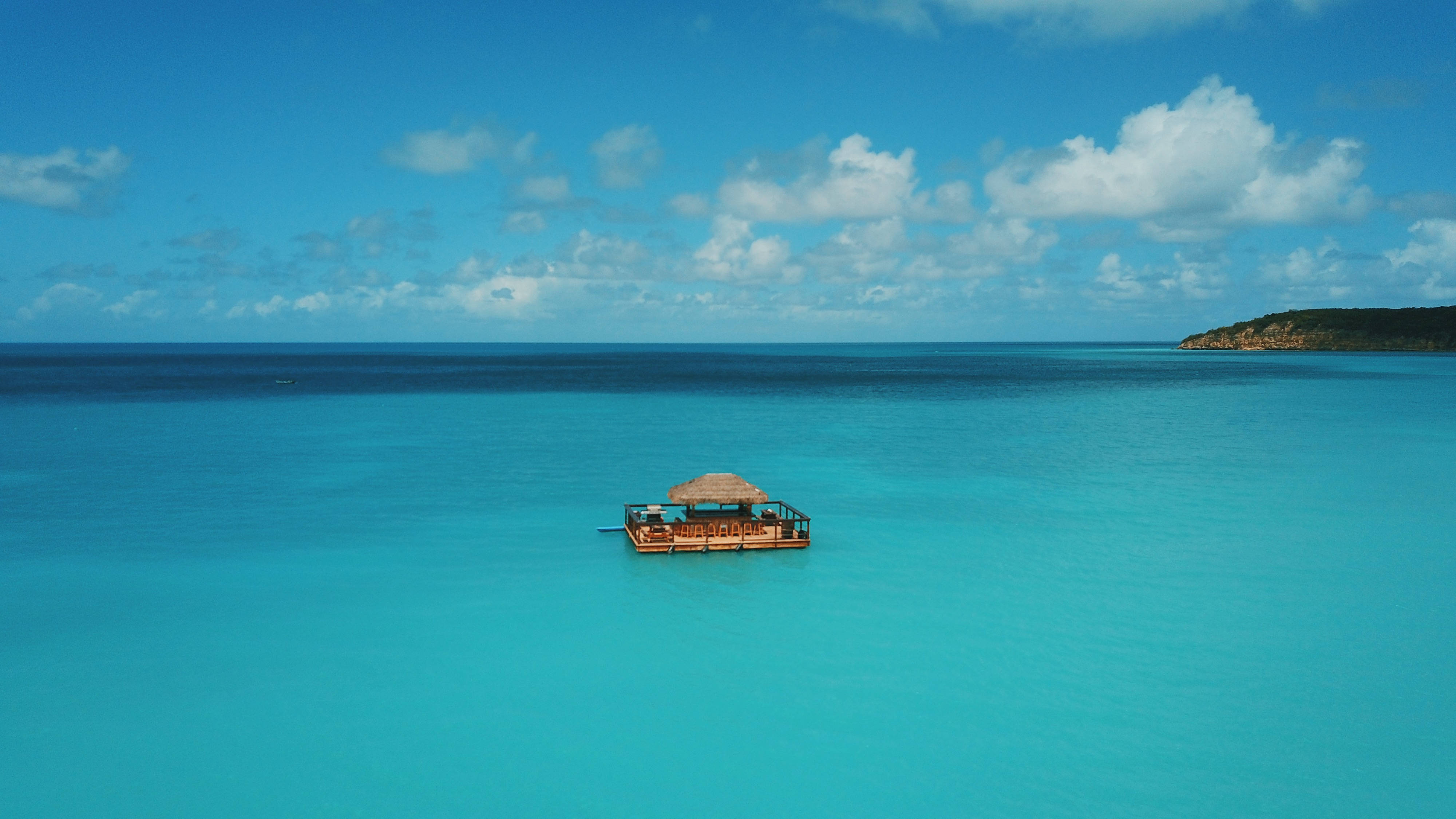 Antigua And Barbuda 桌布 壁紙 — 一個茅草屋頂的木製漂浮酒吧獨自矗立在清澈碧綠的海水中，上方是藍天白雲。