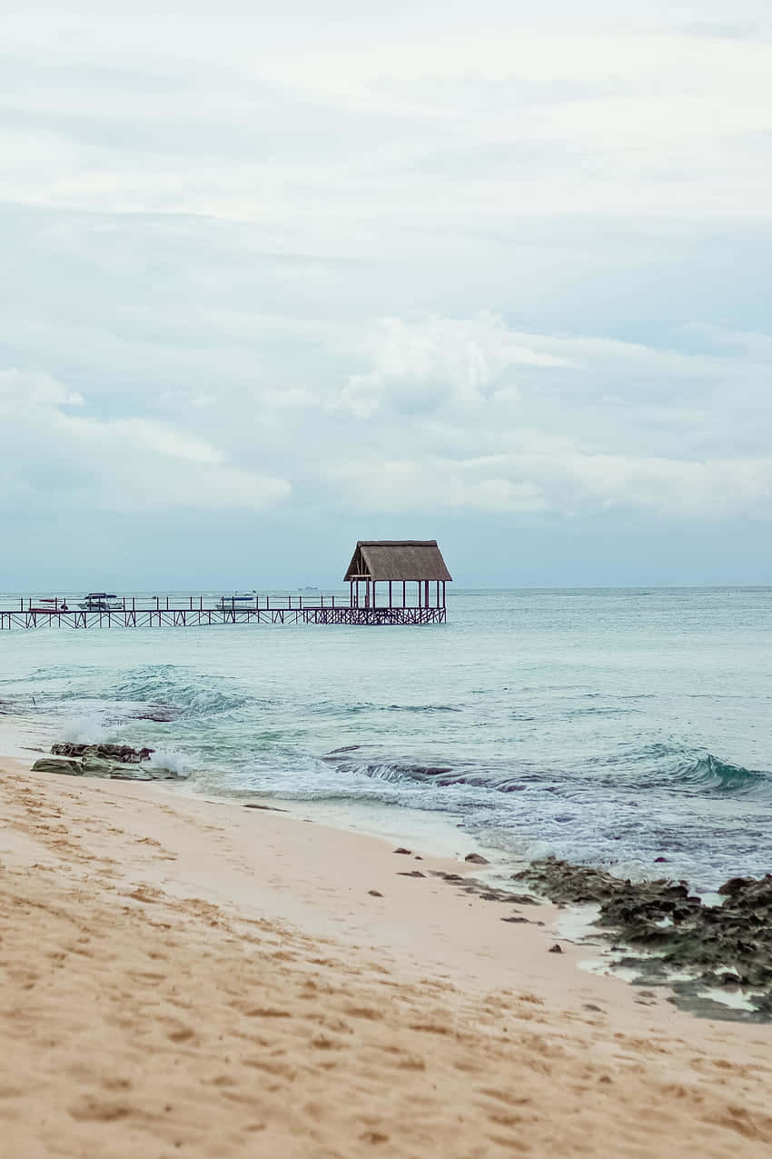 Beach Huts 桌布 壁紙 — 一個茅草屋頂的木製碼頭從沙灘延伸至平靜的碧綠海水，天空多雲。