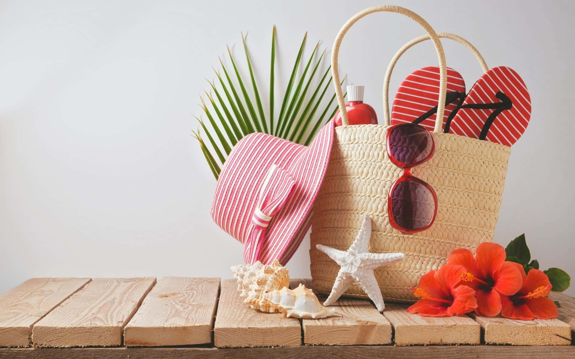 Beach Hat 桌布 壁紙 — 一個草編沙灘袋裝著紅色人字拖、太陽眼鏡和防曬乳，旁邊有粉紅色帽子、海星、貝殼和鮮豔的木芙蓉在木板上。