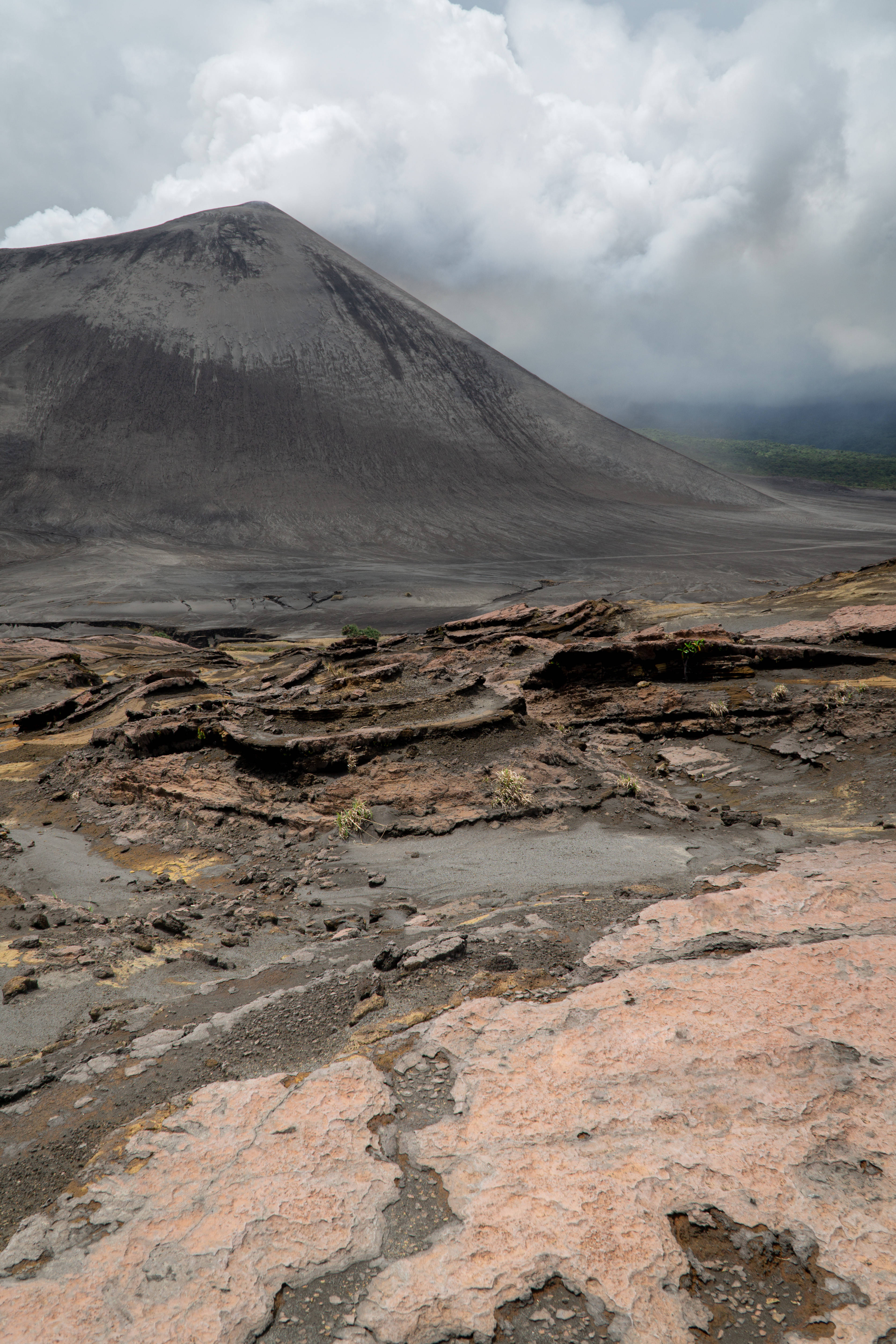 Vanuatu 桌布 壁紙 — 一個荒涼的火山景觀，在陰天之下，巨大的灰色火山錐聳立在龜裂的紅褐色地形後方。