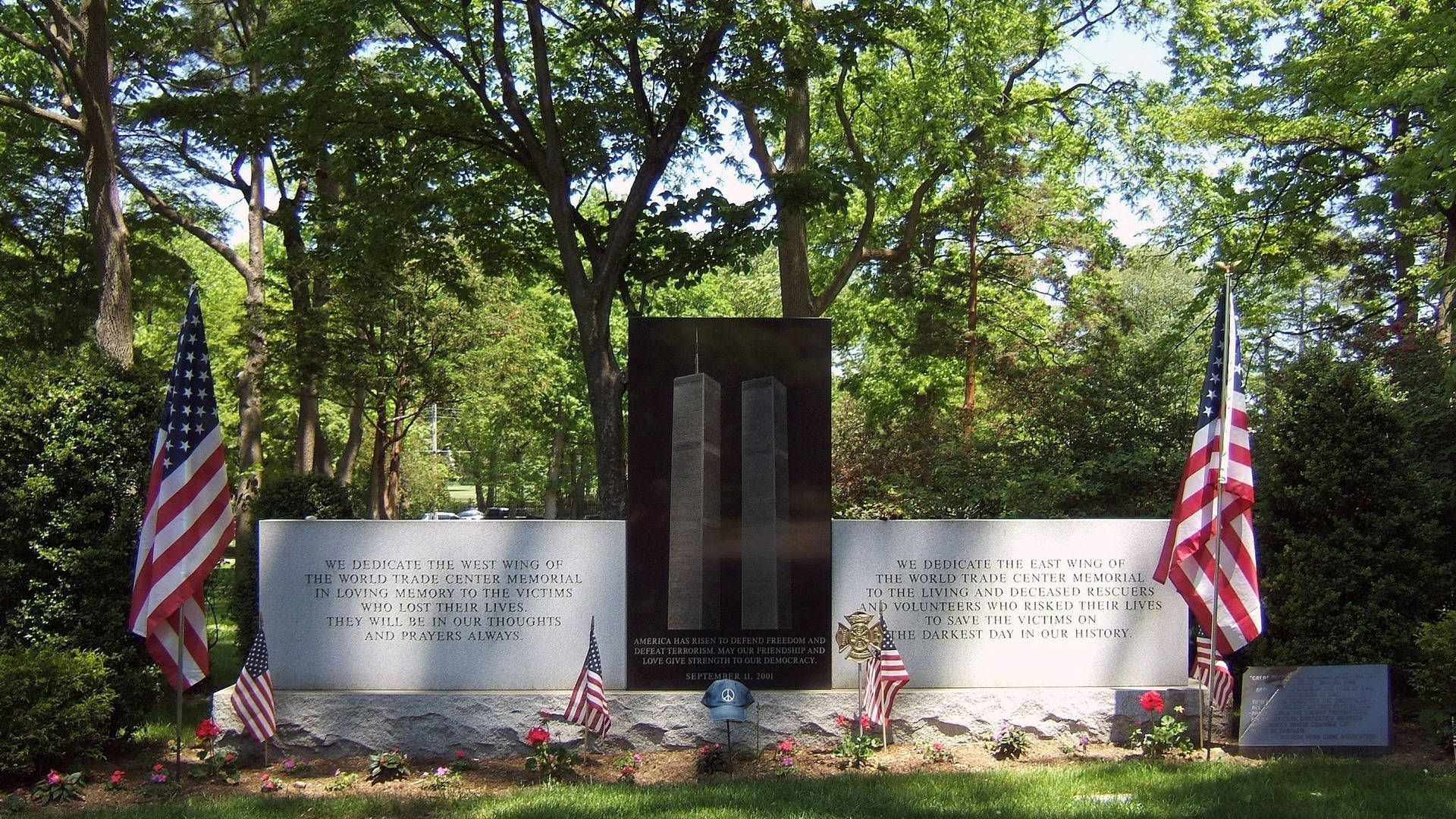 911 Memorial 桌布 壁紙 — 一個莊嚴的世界貿易中心紀念碑，刻有石雕面板和美國國旗，周圍環繞著茂密的綠樹和鮮花。