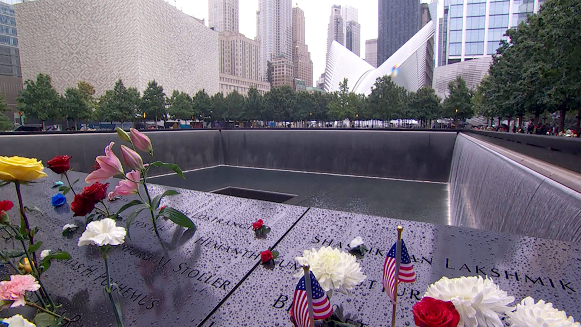 911 Memorial 桌布 壁紙 — 一個莊嚴的紀念池，周圍環繞著層疊的水流和鮮豔的花朵，刻有名字和美國國旗。