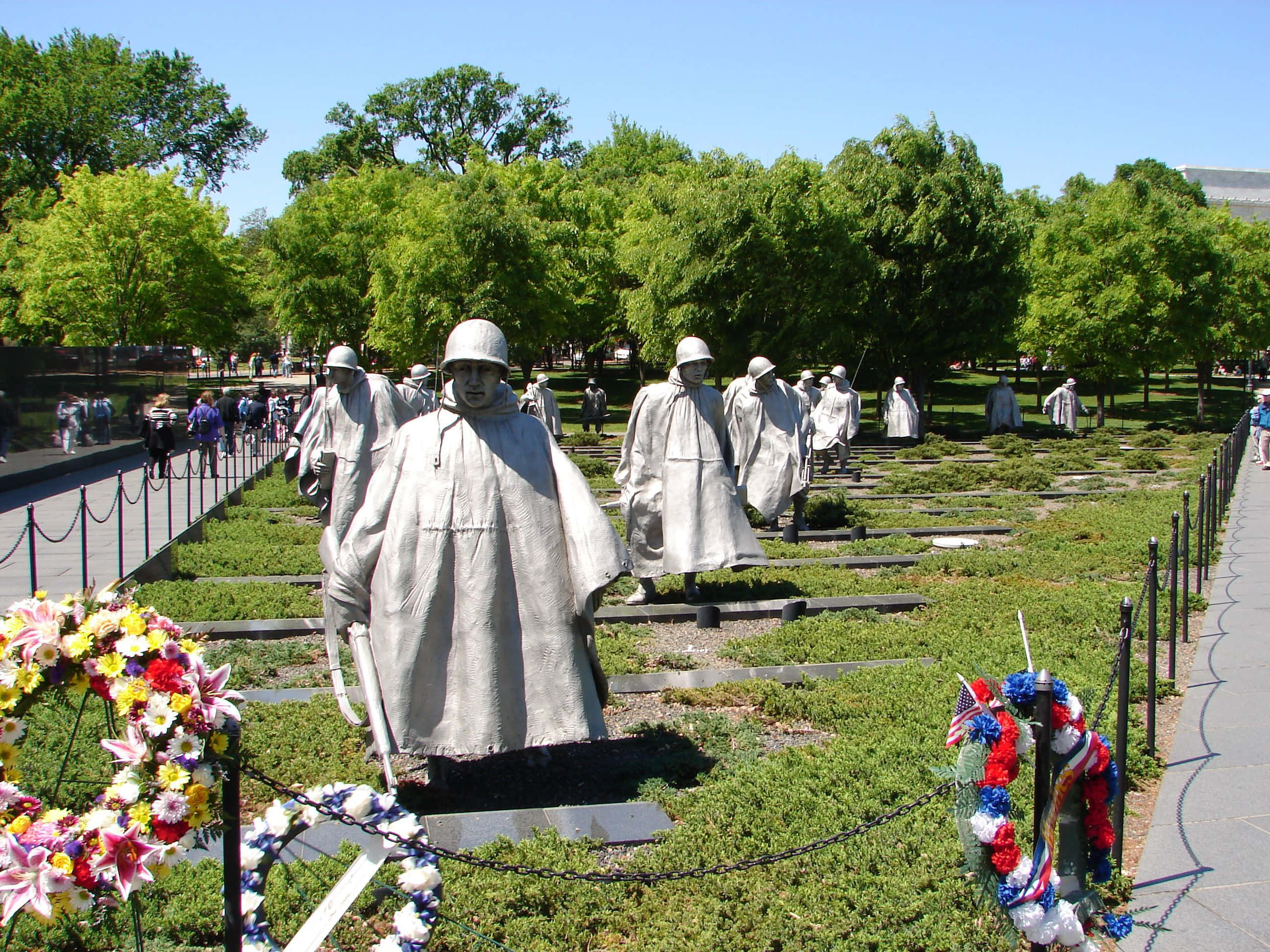 Memorial 桌布 壁紙 — 一個莊嚴的紀念碑，灰色士兵雕像在晴朗的藍天下穿過草地公園，底座上擺放著色彩繽紛的花圈。