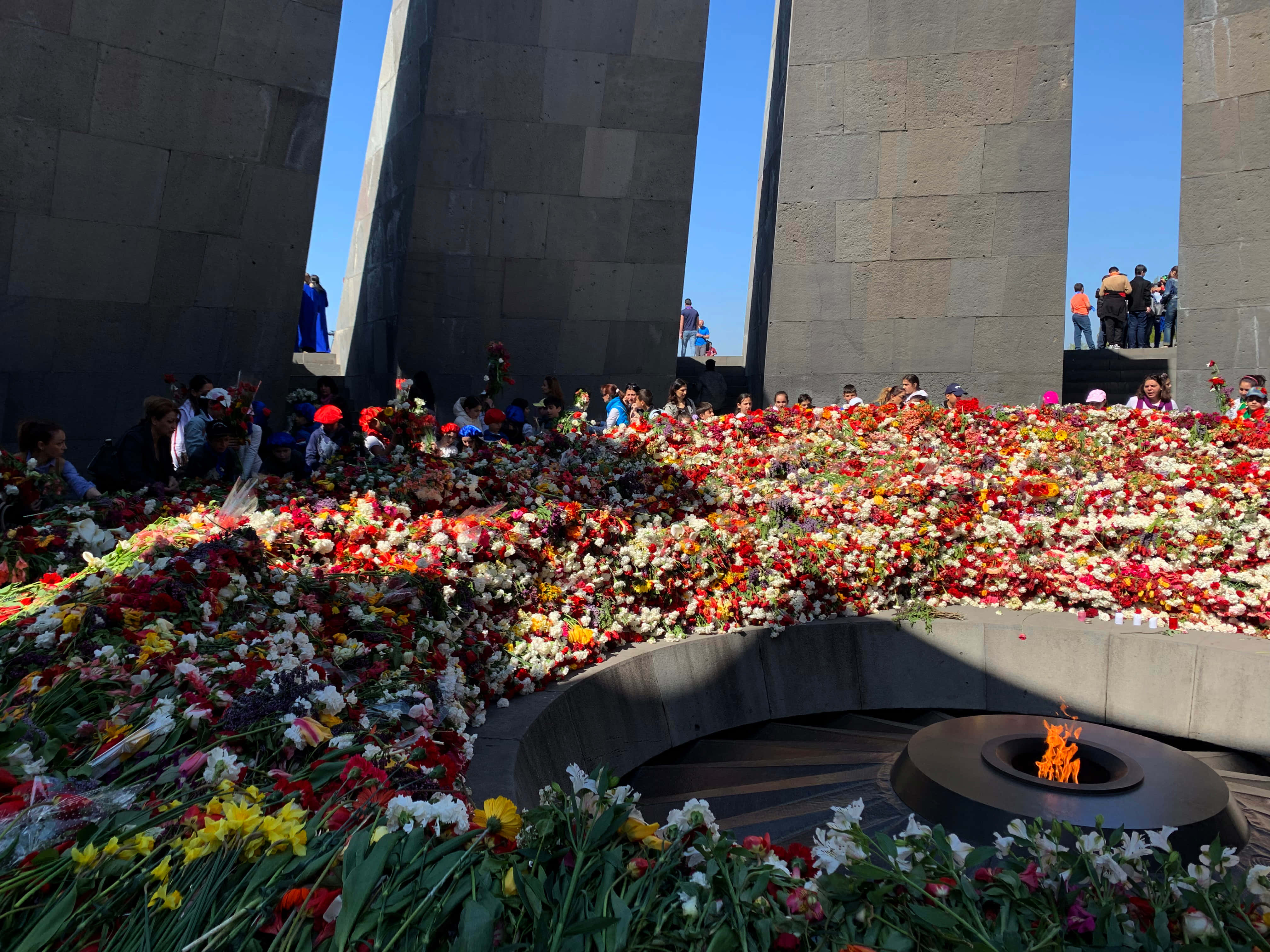 Memorial 桌布 壁紙 — 一個莊嚴的紀念遺址，高聳的石柱和紅、白、黃色花海環繞著永恆之火，在晴朗的藍天下。