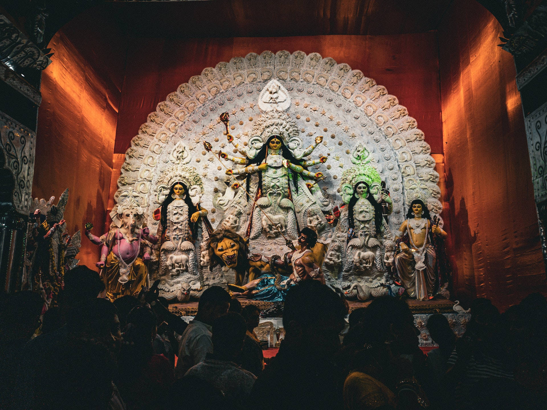 Hindu Temple 桌布 壁紙 — 一個華麗宏偉的杜爾加普祭壇，有多個女神雕像，被溫暖的橙色燈光照亮，從一群崇拜者背後觀看。
