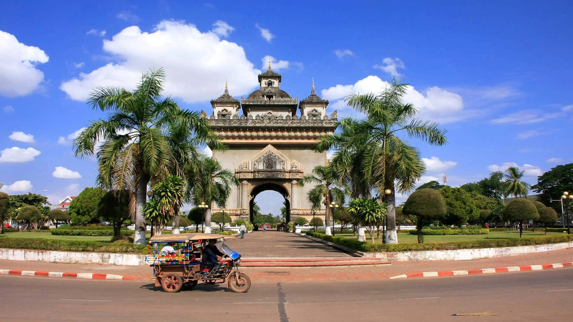 Laos 桌布 壁紙 — 一個華麗的大型拱門矗立在晴朗的藍天白雲下，兩旁種有棕櫚樹，一輛機動三輪車駛過。