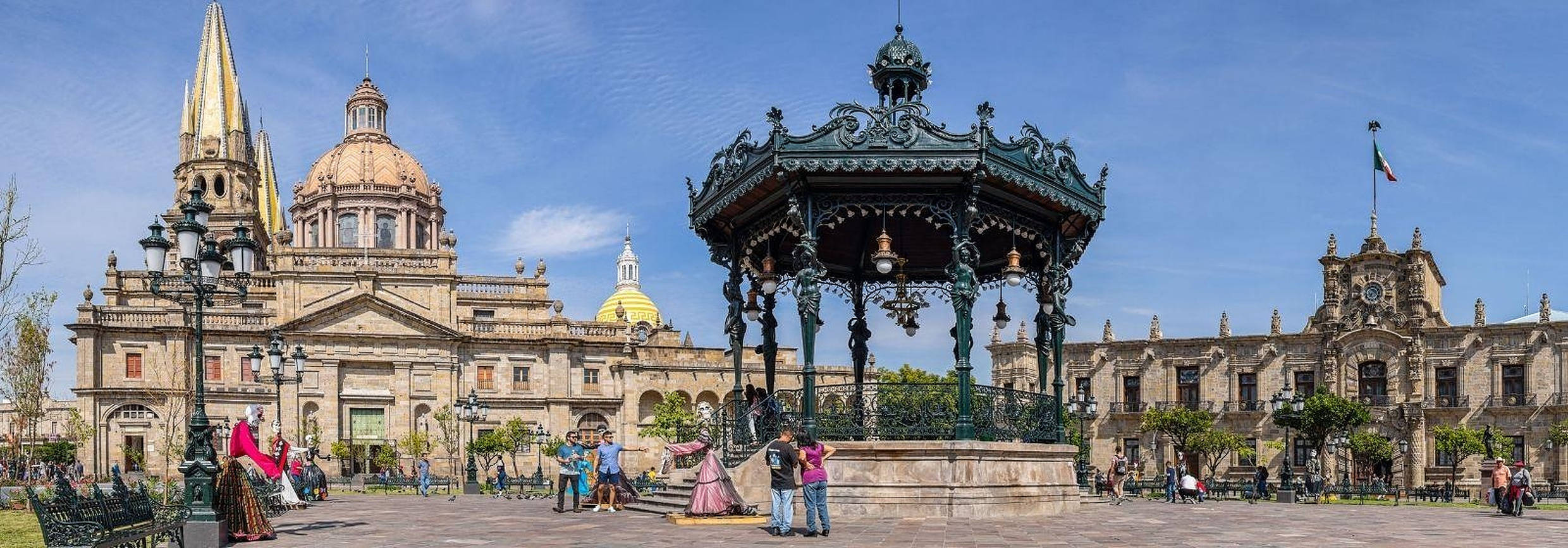 Guadalajara 桌布 壁紙 — 一個華麗的大型涼亭矗立在陽光普照的廣場上，兩旁是歷史悠久的新古典主義建築，上方是清澈的藍天。