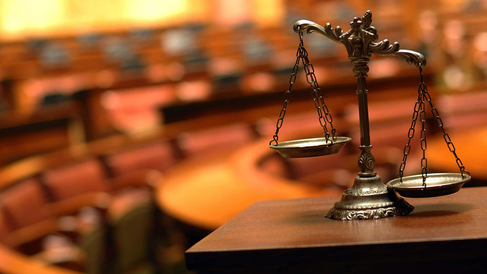 Court 桌布 壁紙 — 一個華麗的金屬正義天平平衡地放在一張木桌上，背景是模糊的法庭，有一排排空座位。