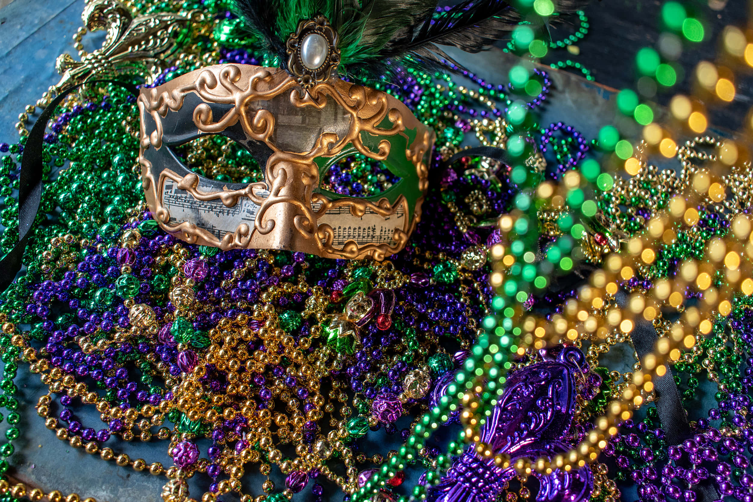 Mardi Gras 桌布 壁紙 — 一個華麗的金色狂歡節面具放在一堆充滿活力的綠色、紫色和金色珠子上，置於質樸的木質表面上。