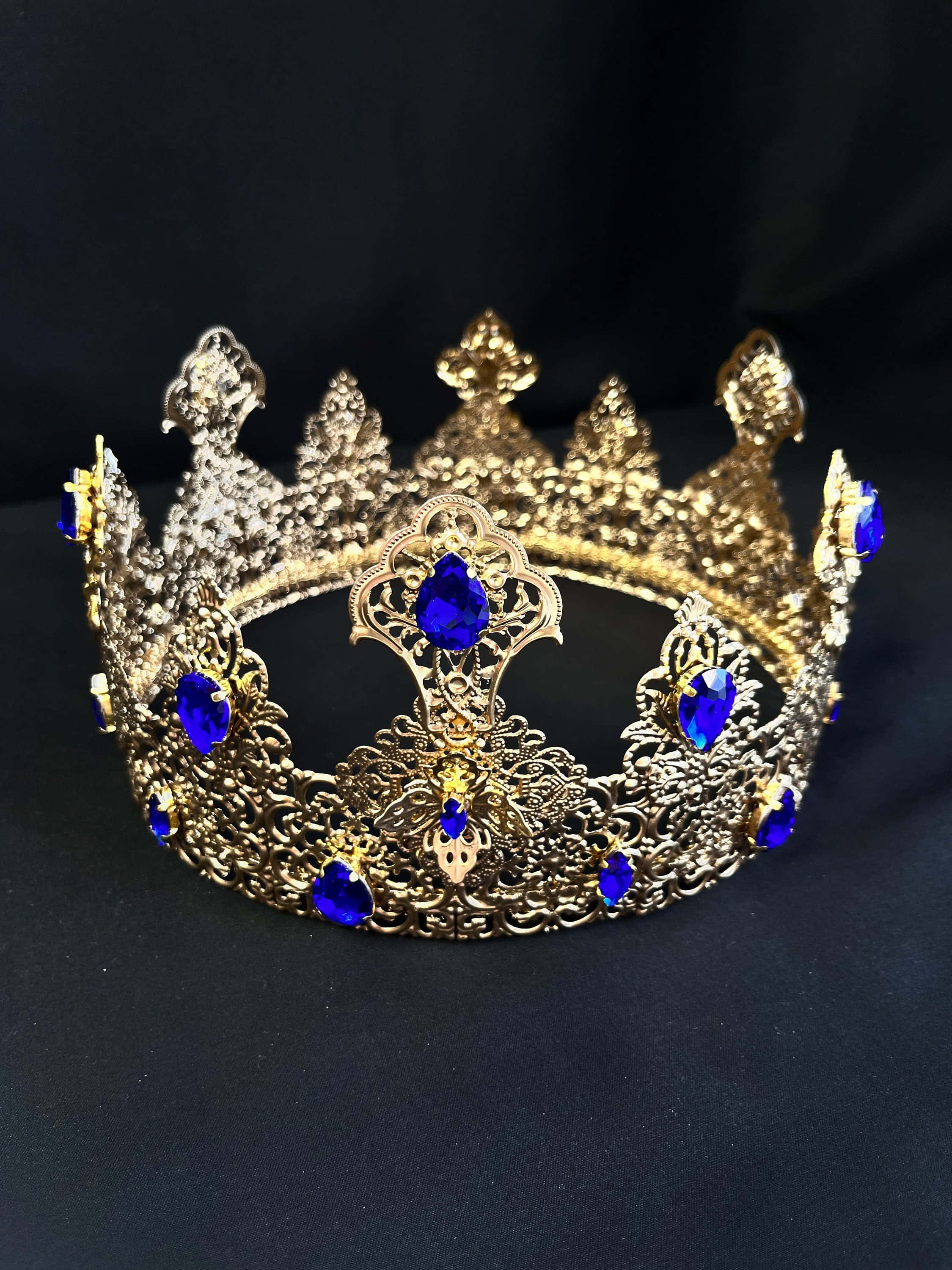 Crown 桌布 壁紙 — 一個華麗的金色皇冠，帶有精緻的鏤空花紋和深藍色寶石，靠在深色布料背景上。
