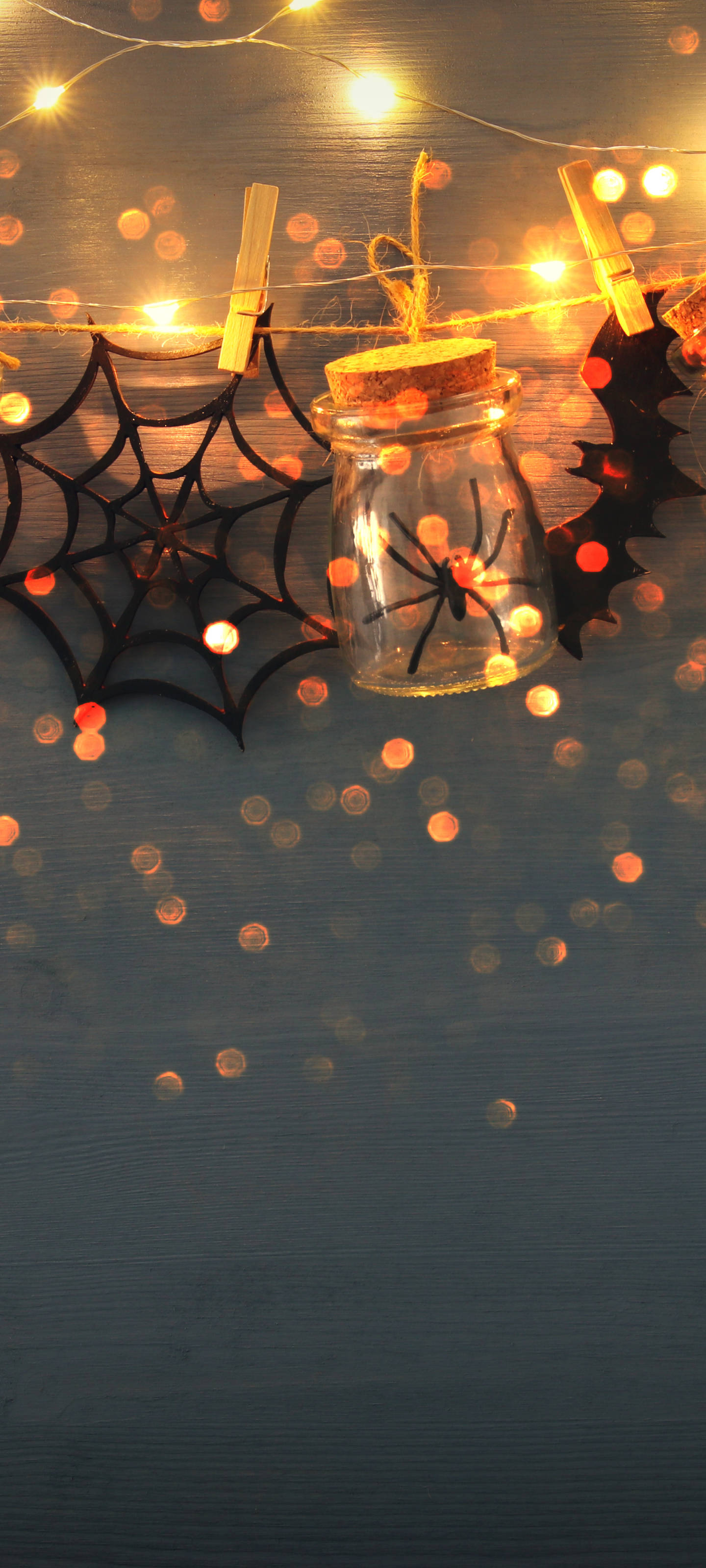Halloween Phone 桌布 壁紙 — 一個萬聖節主題的罐子，裡面有一隻黑色蜘蛛，放在藍色木質表面上，周圍環繞著燈串、蜘蛛網和節日裝飾。