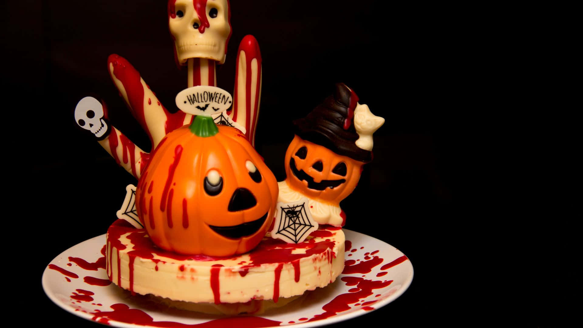 Halloween Cake 桌布 壁紙 — 一個萬聖節主題的起司蛋糕，上面有微笑的南瓜、殭屍手和骷髏裝飾，背景為黑色。