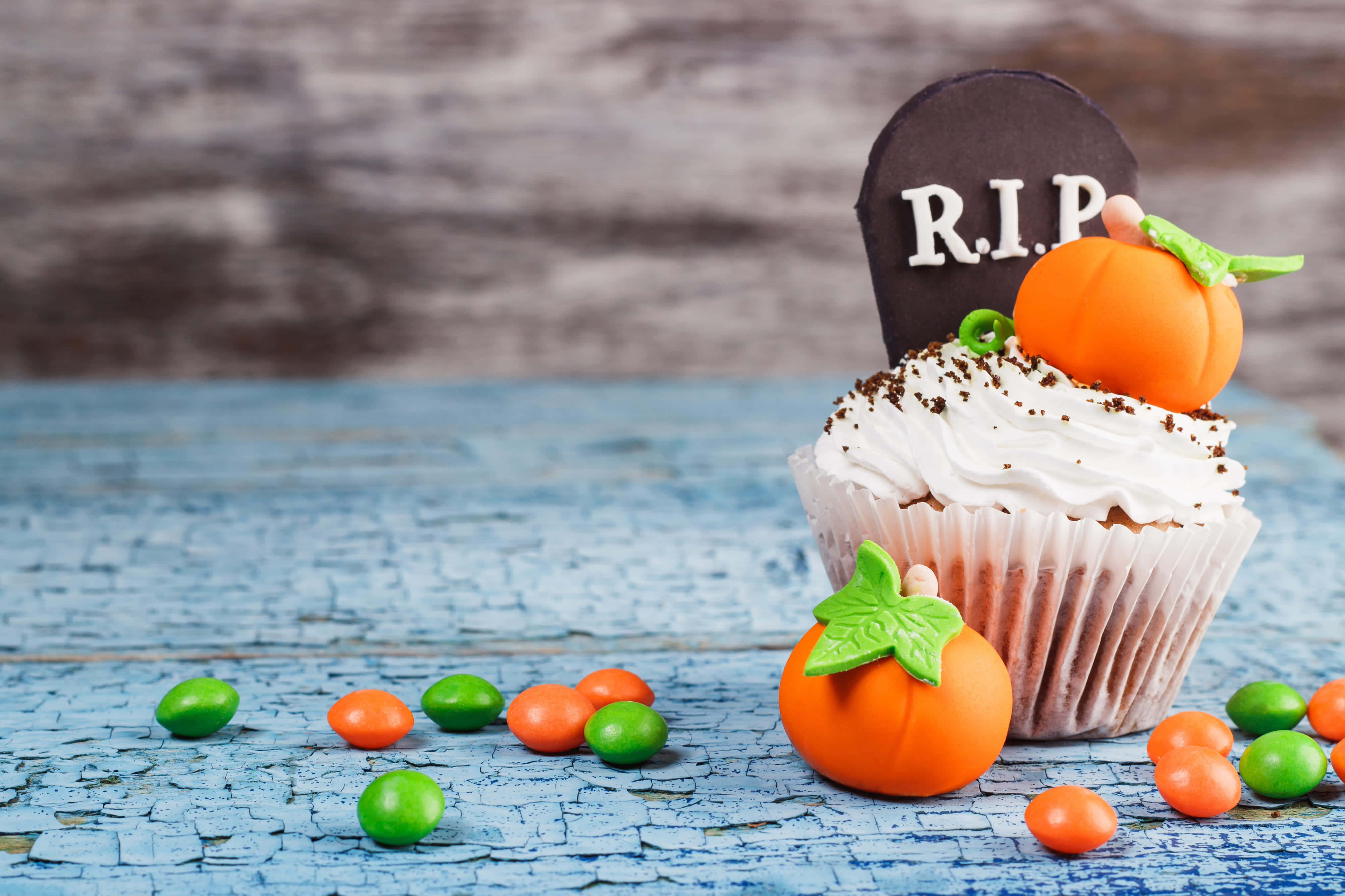 Halloween Cupcakes 桌布 壁紙 — 一個萬聖節杯子蛋糕，上面有墓碑和南瓜裝飾，放在一個風化的藍色木質表面上，周圍環繞著糖果。