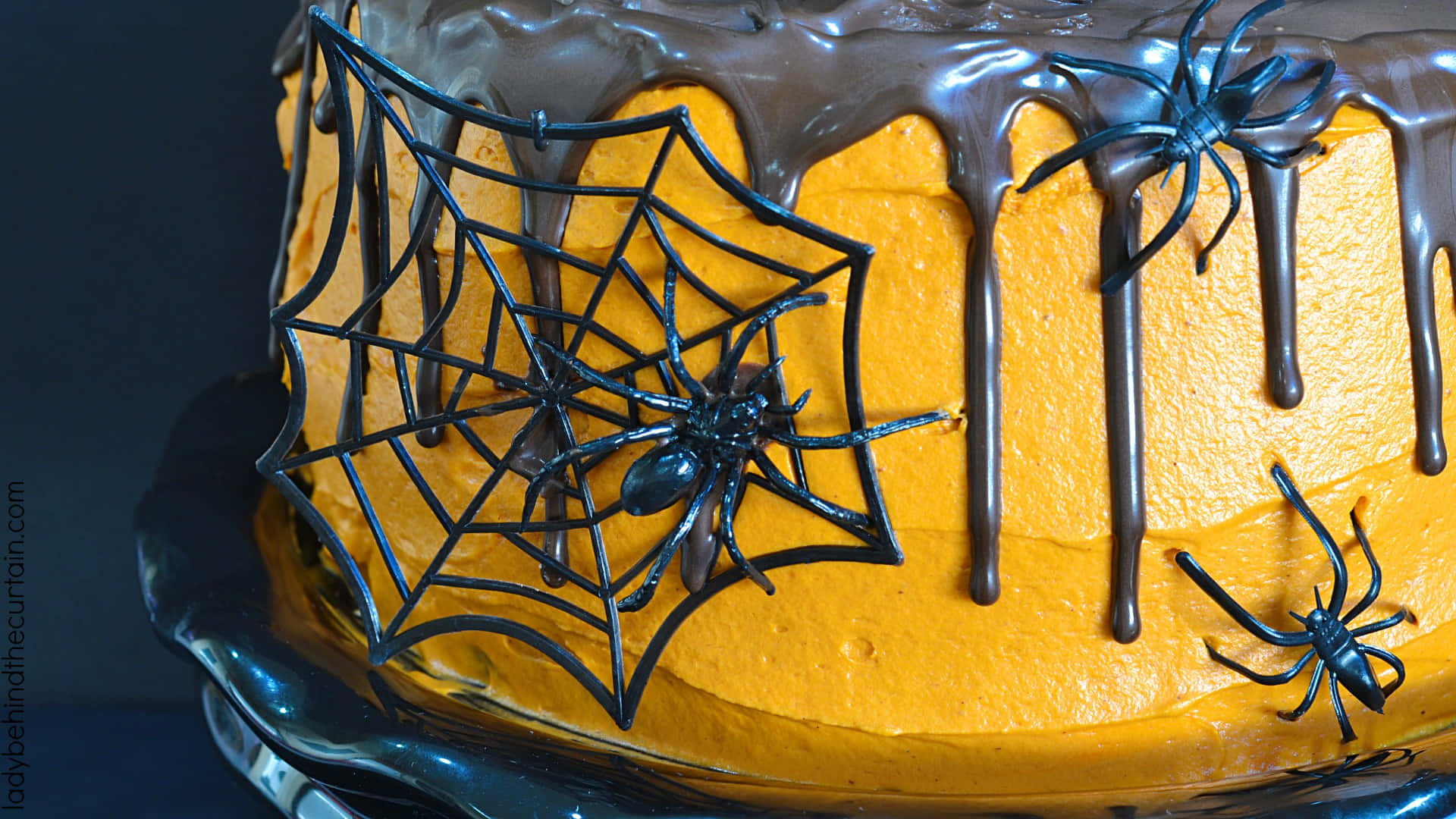 Halloween Cake 桌布 壁紙 — 一個萬聖節蛋糕，帶有橙色糖霜、巧克力滴和蜘蛛網上的黑色塑膠蜘蛛。