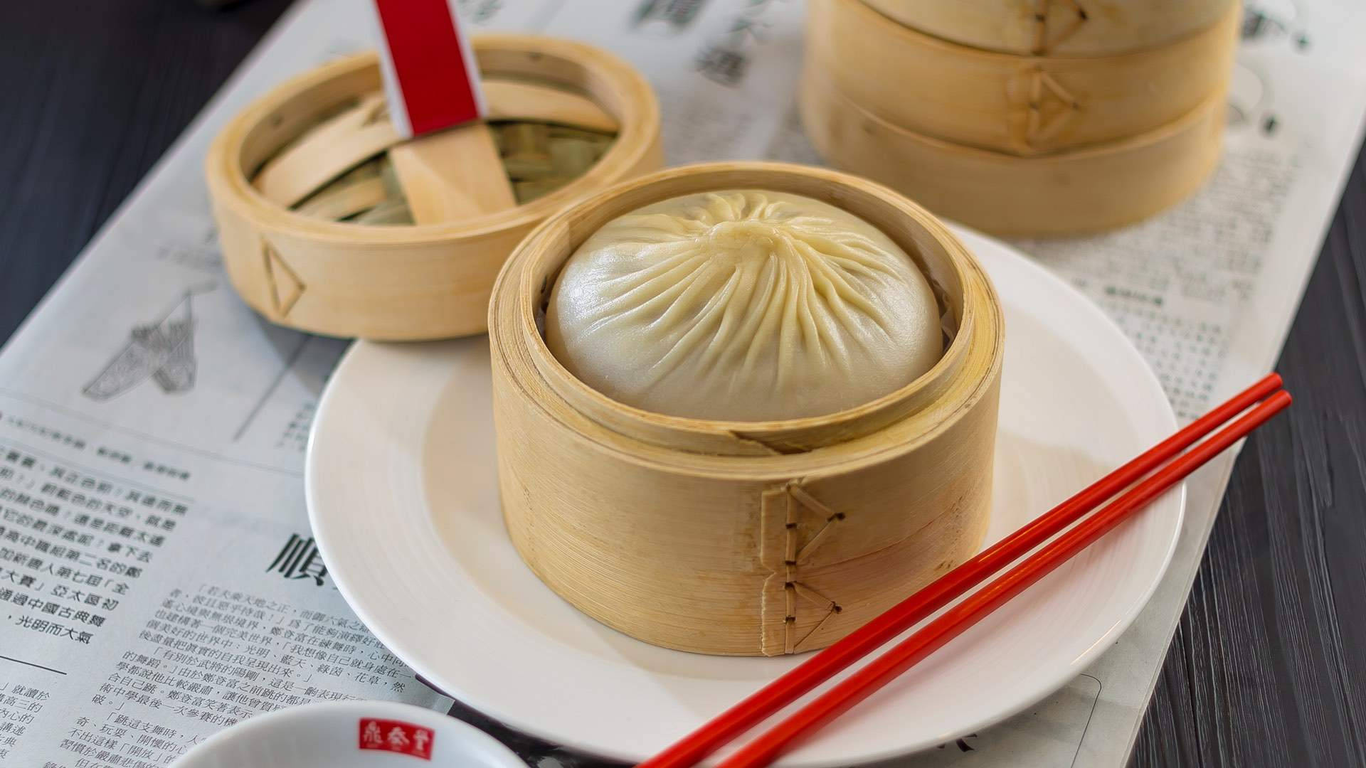 steam 桌布 壁紙 — 一個蒸餃完美地放在竹蒸籠裡，放在報紙上，旁邊有紅色筷子。