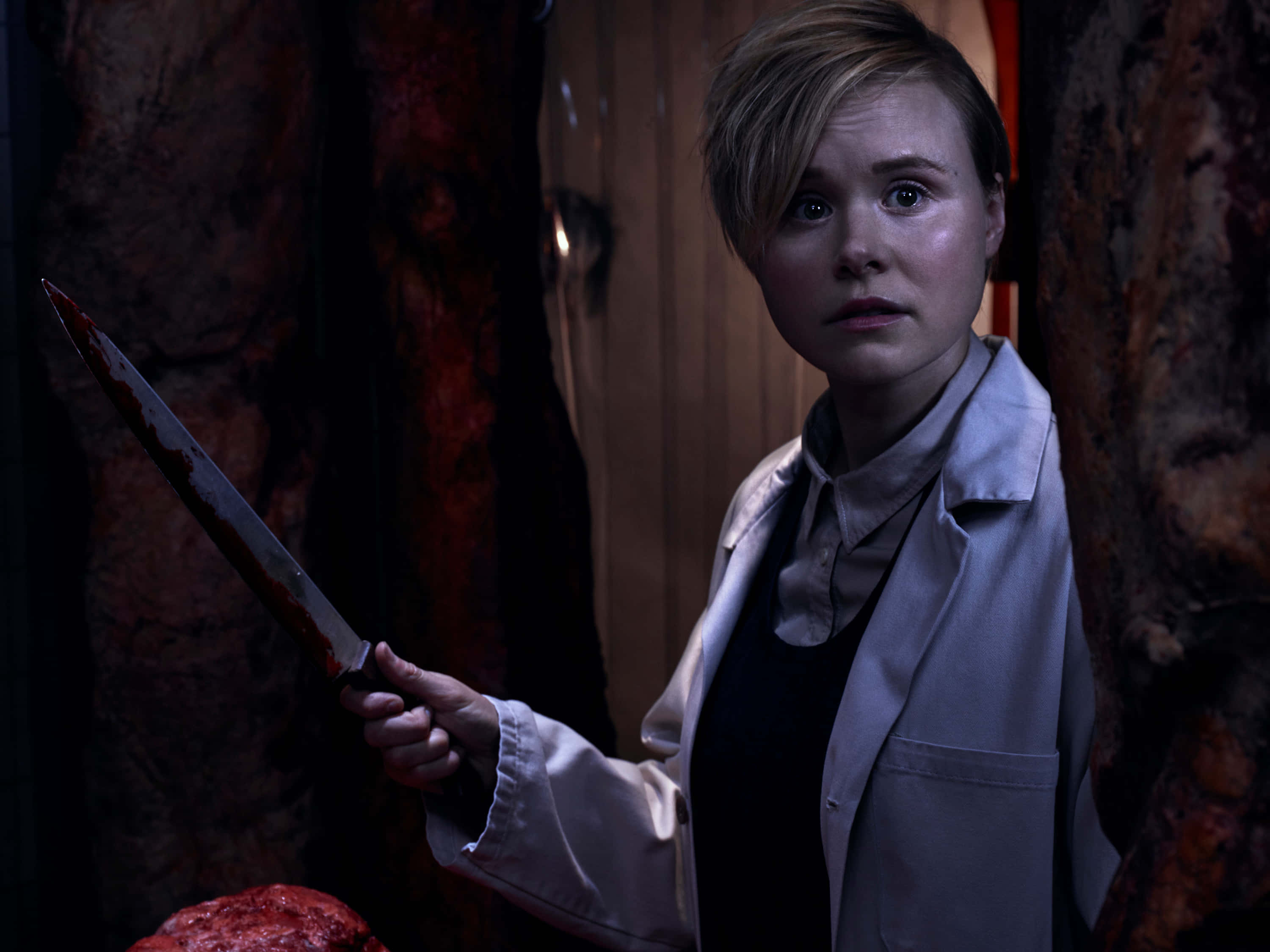 Alison Pill 桌布 壁紙 — 一個蒼白的女人穿著白色實驗室外套，在一個光線昏暗、掛著肉的房間裡拿著一把血淋淋的刀。