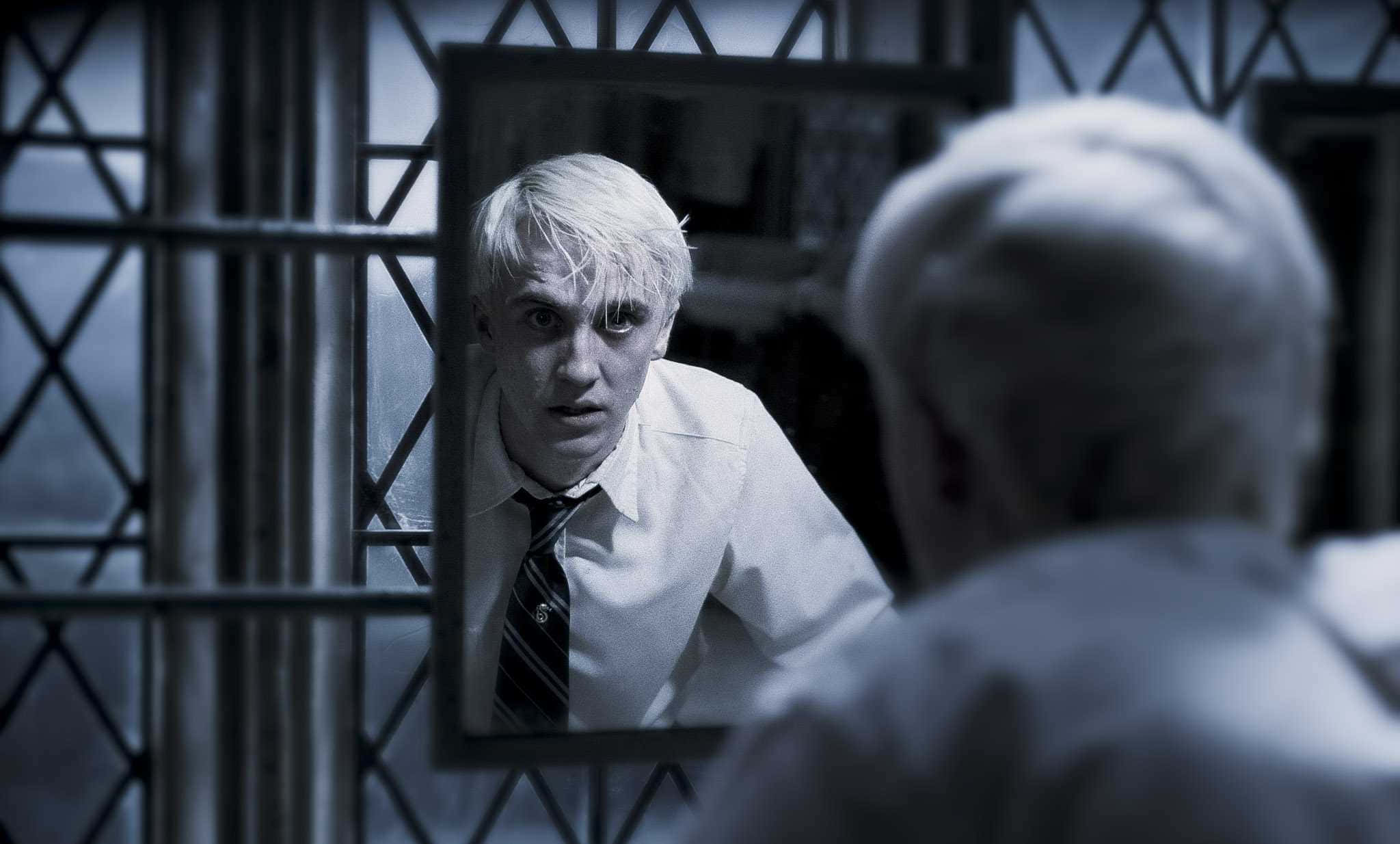 Tom Felton 桌布 壁紙 — 一個蒼白的年輕男子，身穿白襯衫和條紋領帶，在昏暗的房間裡，隔著鐵欄杆，凝視著鏡中的自己。