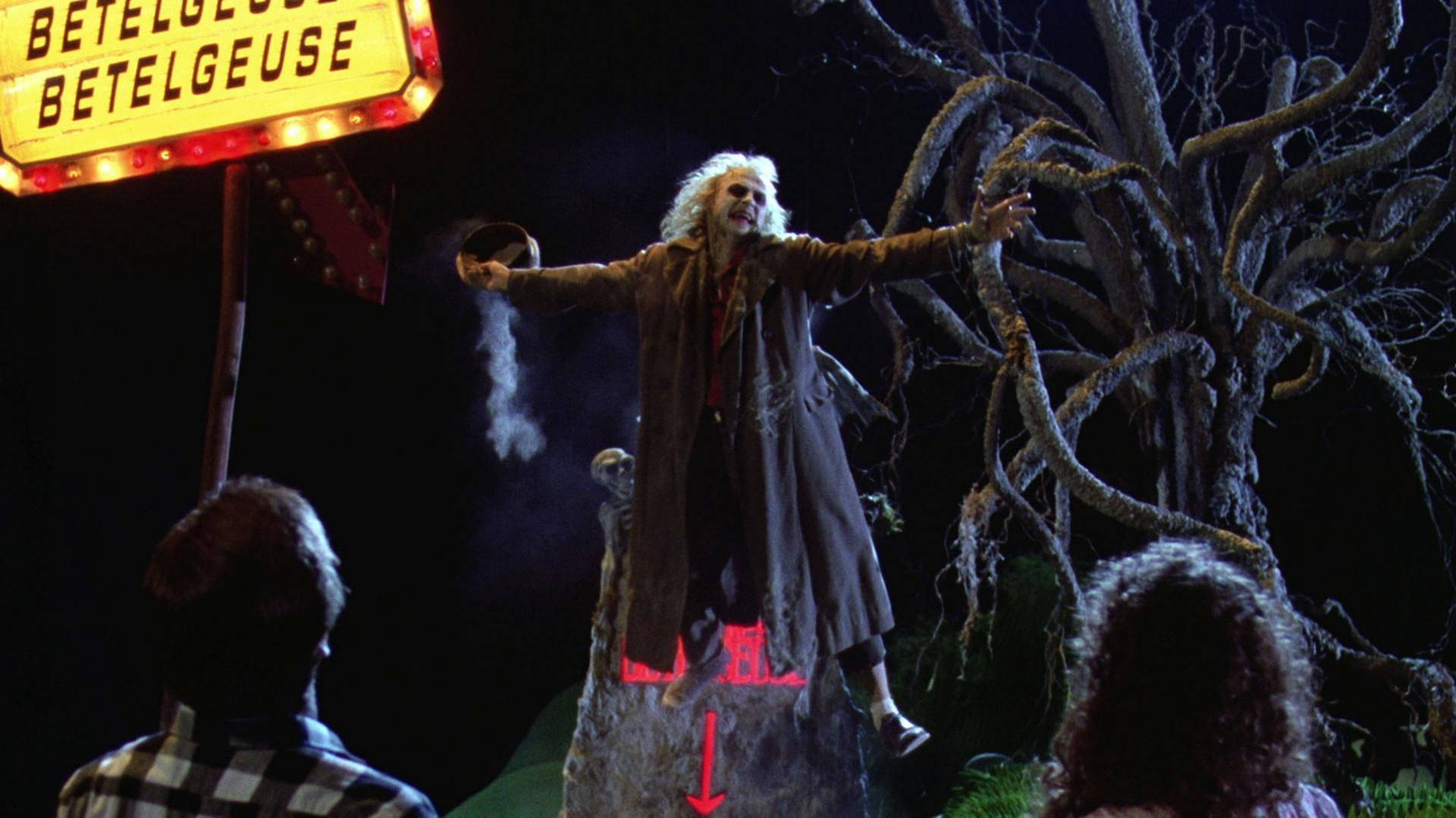 Beetlejuice 桌布 壁紙 — 一個蒼白的男人頭髮凌亂，在夜晚站在發光的「BETELGEUSE」標誌下的岩石上，在扭曲的樹木前伸展雙臂。