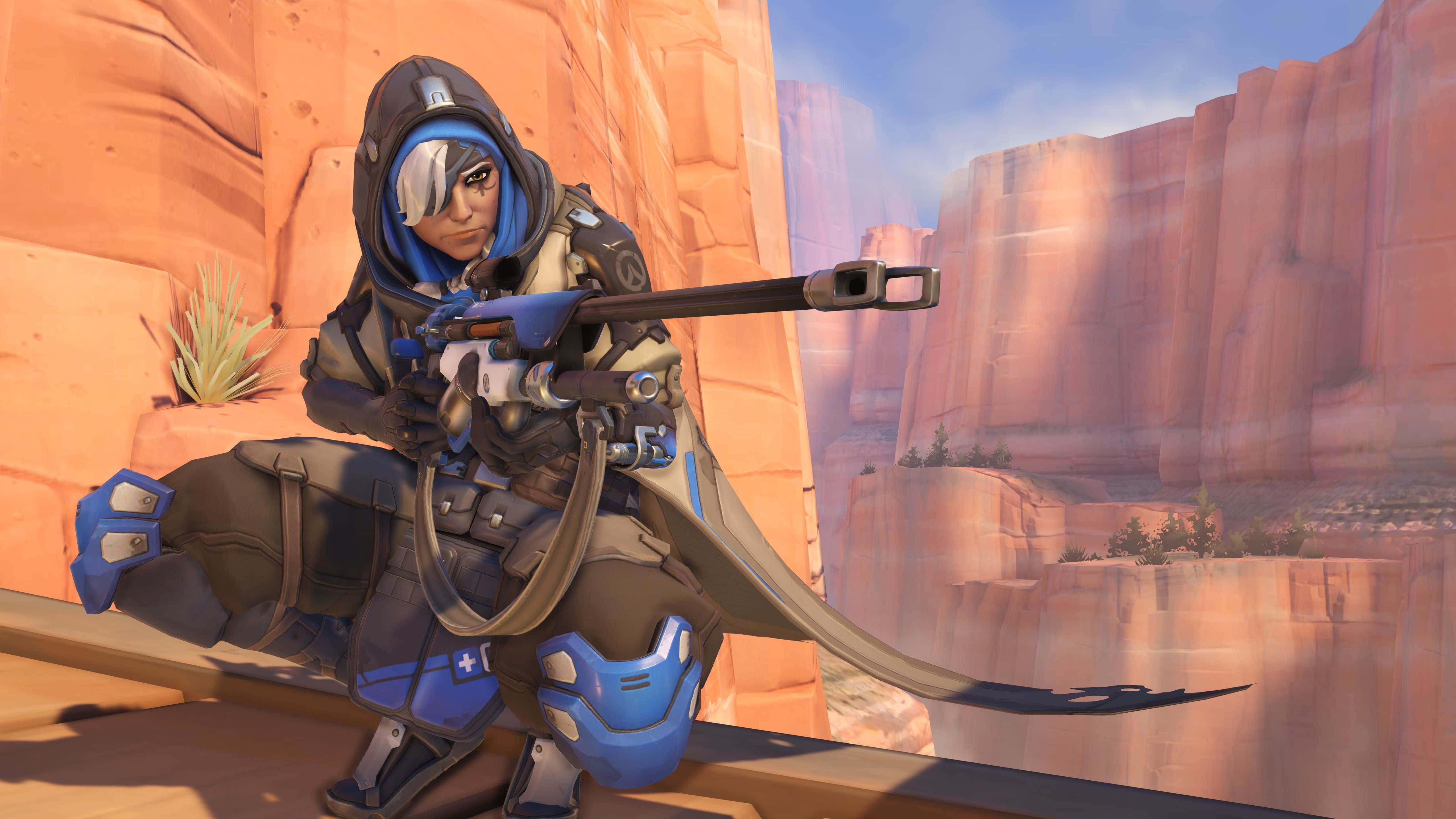 Overwatch Ana 桌布 壁紙 — 一個藍灰色裝甲角色蹲在壁架上，瞄準狙擊步槍，背景是晴朗藍天下高聳的橙色岩層。