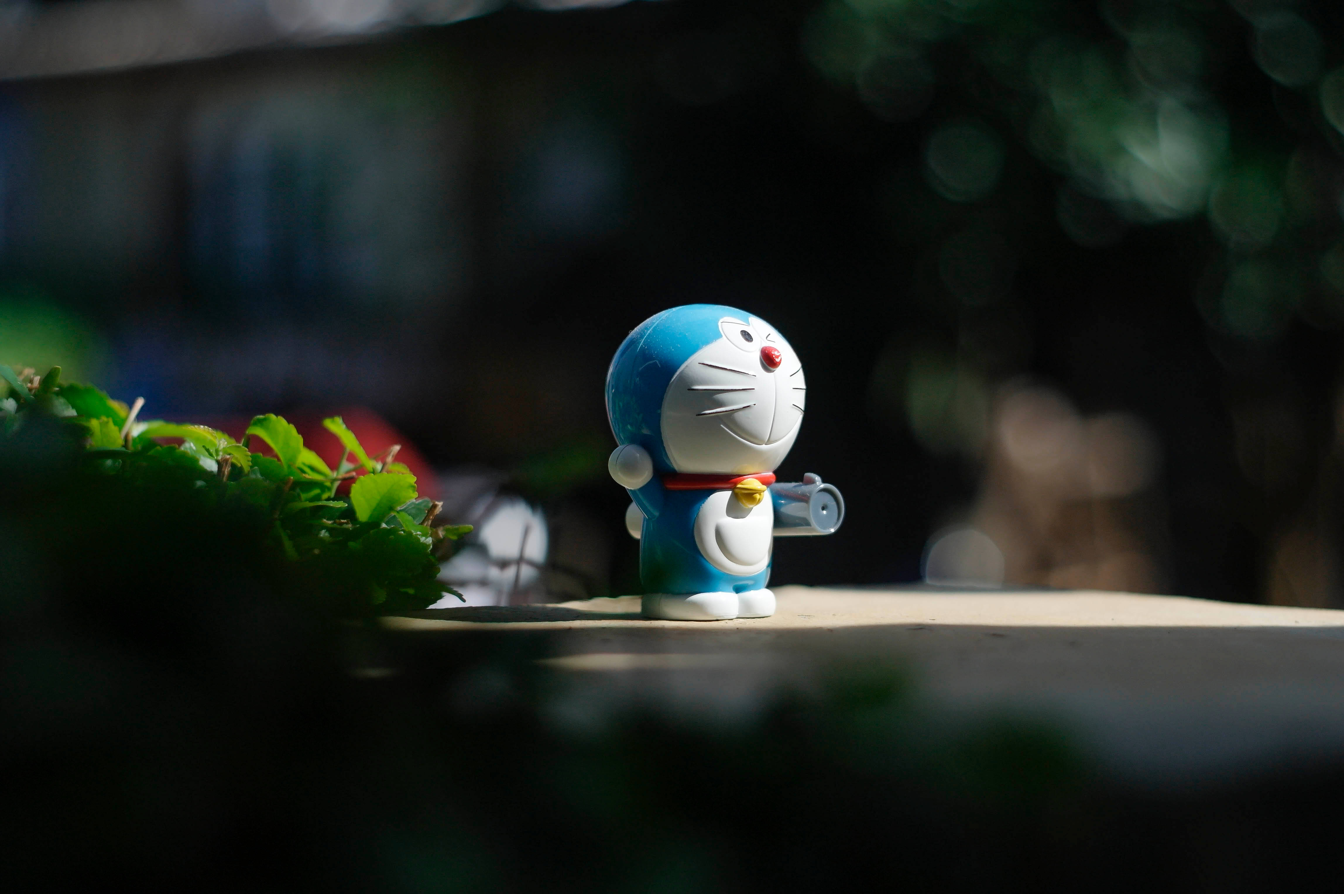 Doraemon 桌布 壁紙 — 一個藍白色的哆啦A夢公仔站在柔和陽光下，旁邊有綠色樹葉，背景是模糊的戶外景色。