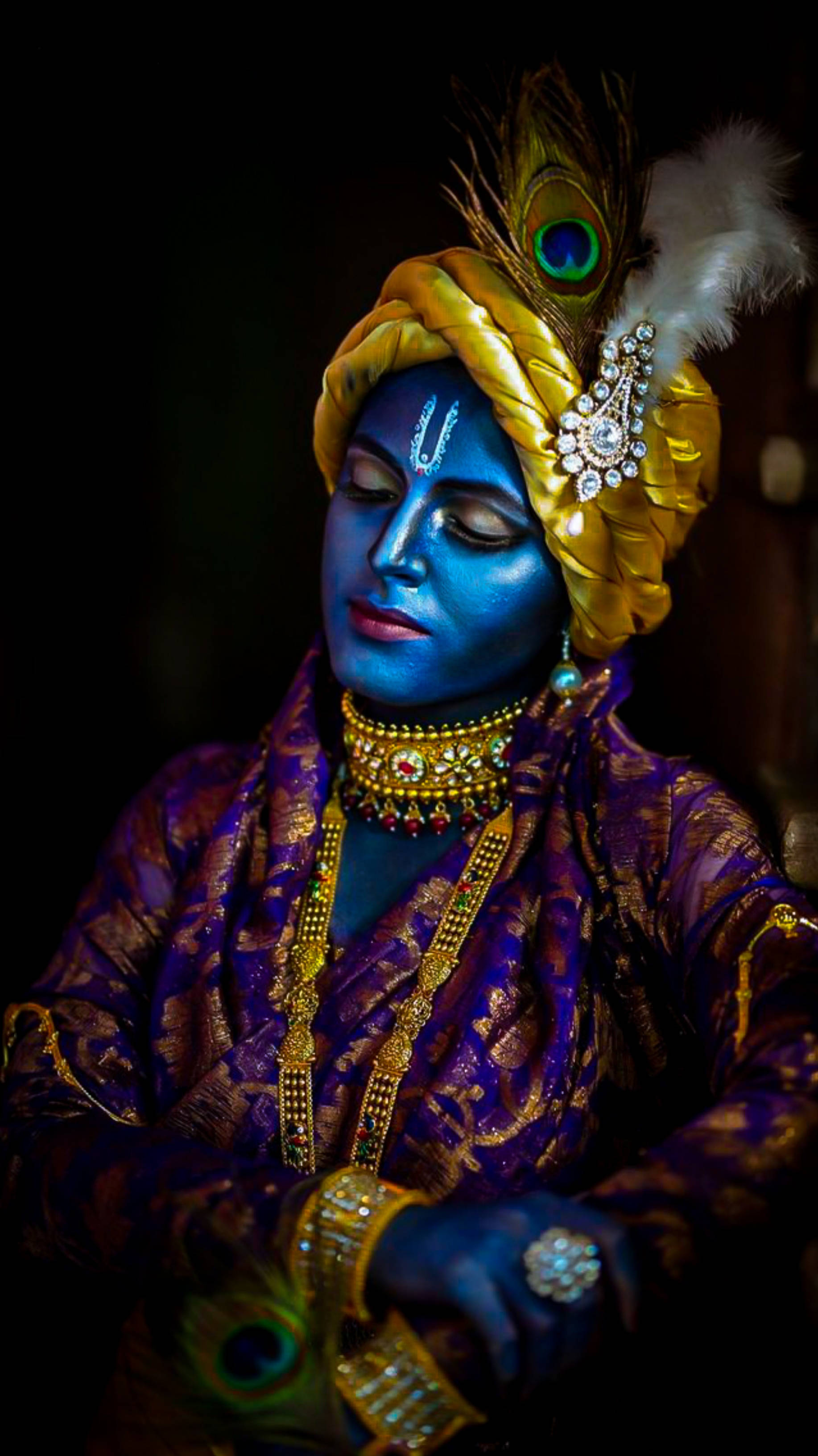 krishna iPhone 桌布 壁紙 — 一個藍皮膚、戴著飾有孔雀羽毛的金色頭巾的人，穿著華麗的紫色和金色服裝，靜靜地凝視著。