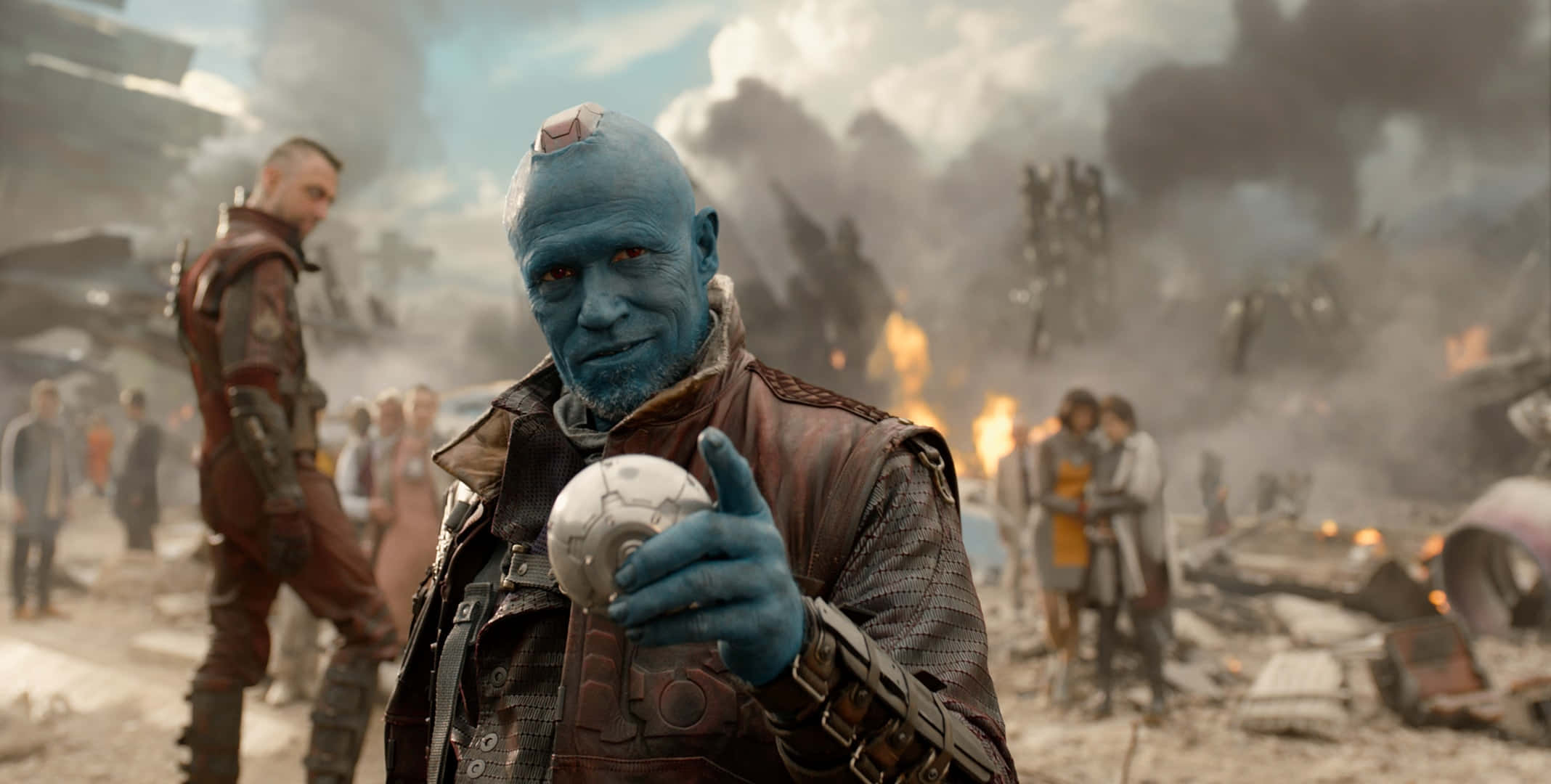 Guardians Of The Galaxy 2 桌布 壁紙 — 一個藍皮膚、紅眼睛的外星人在煙霧彌漫的戰場上手持發光球體，背景有燃燒的殘骸和士兵。