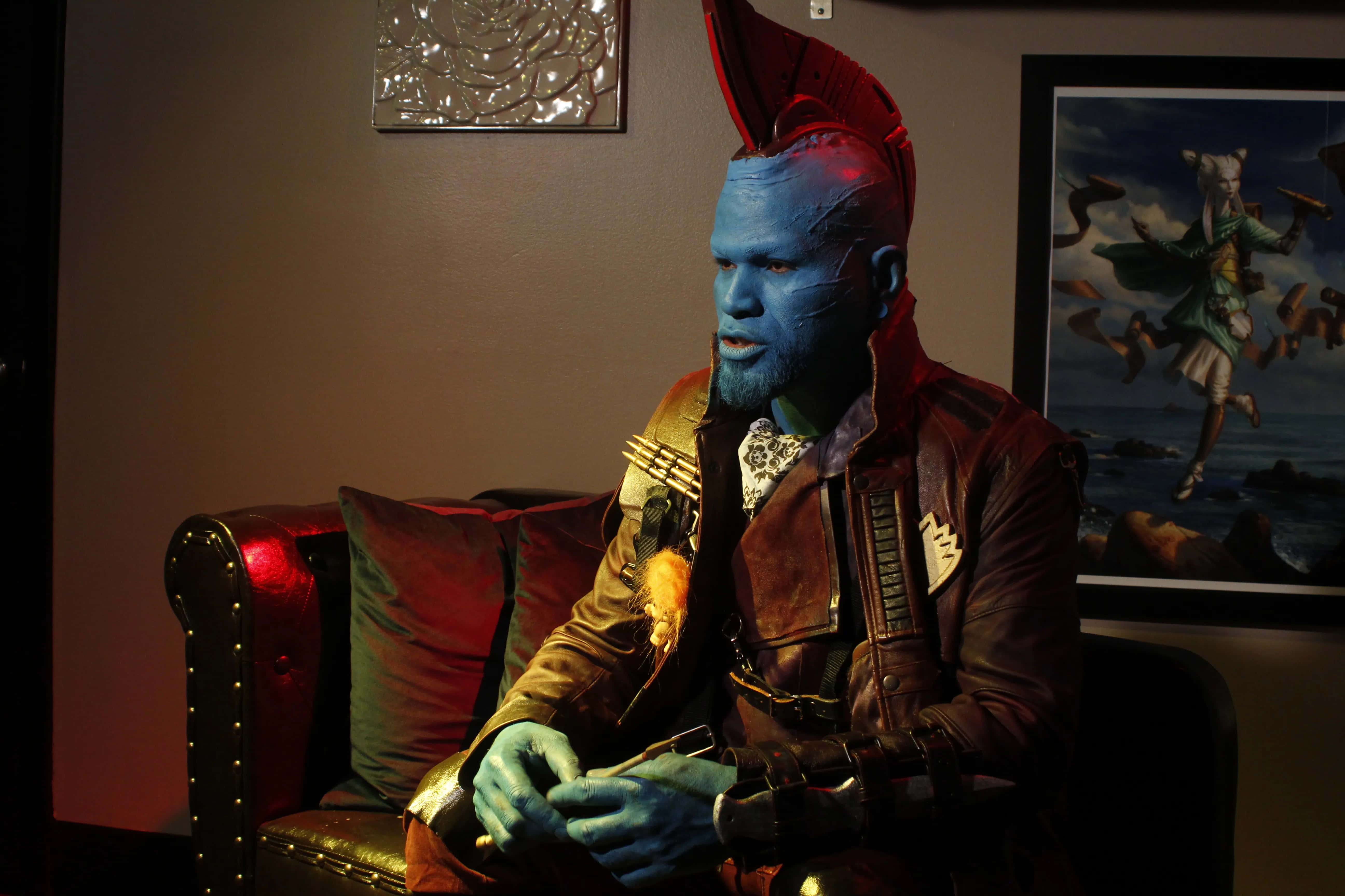 Yondu 桌布 壁紙 — 一個藍皮膚、紅色莫西干頭的外星人坐在皮椅上，在昏暗的紅光下拿著槍。