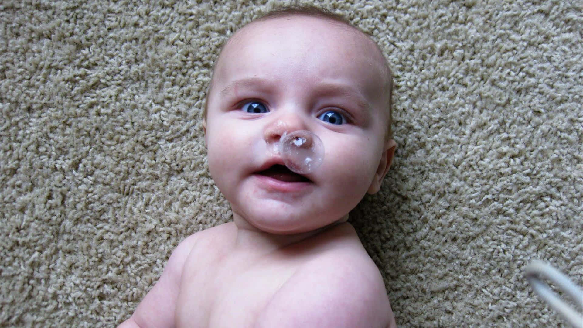 Funny Baby 桌布 壁紙 — 一個藍眼睛、戴著鼻導管的嬰兒躺在米色地毯上，好奇地向上看。