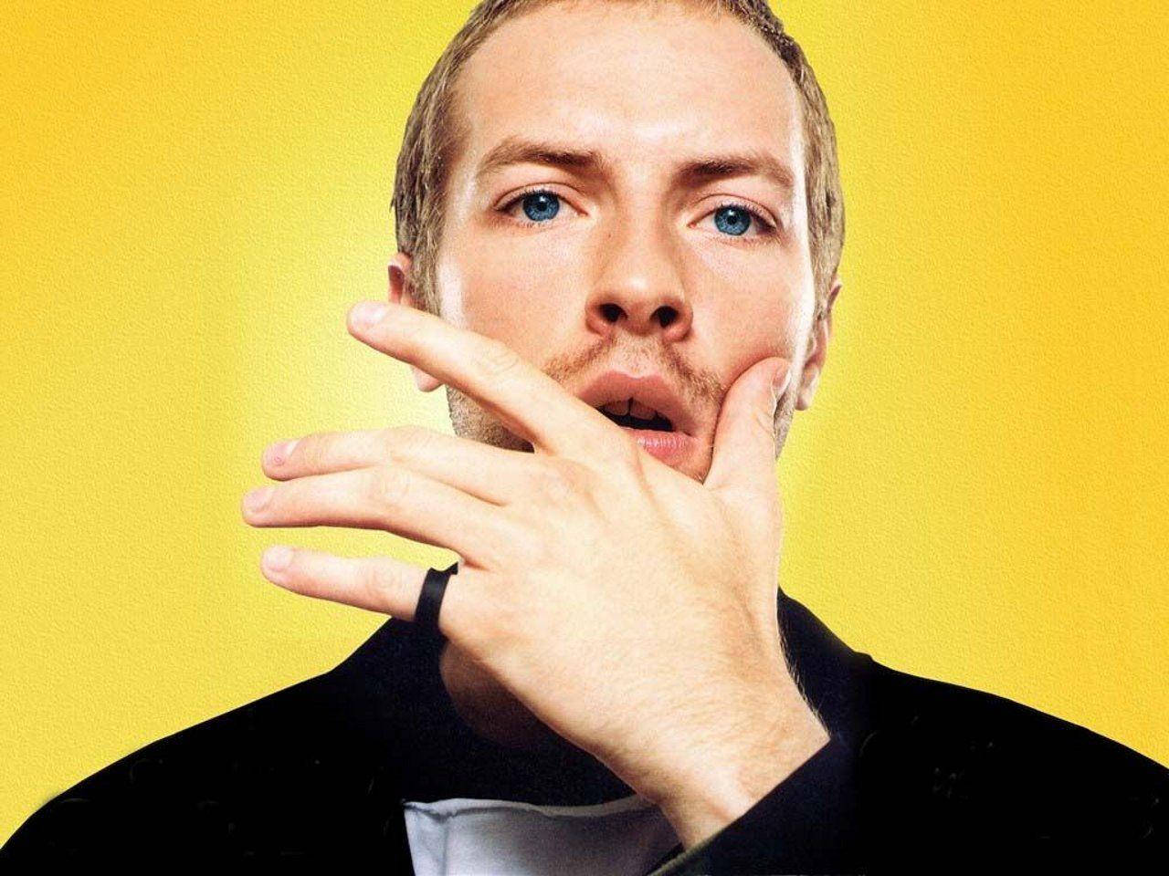 Chris Martin 桌布 壁紙 — 一個藍眼睛、留鬍鬚的男人，在明亮的黃色背景前將手靠近臉部。
