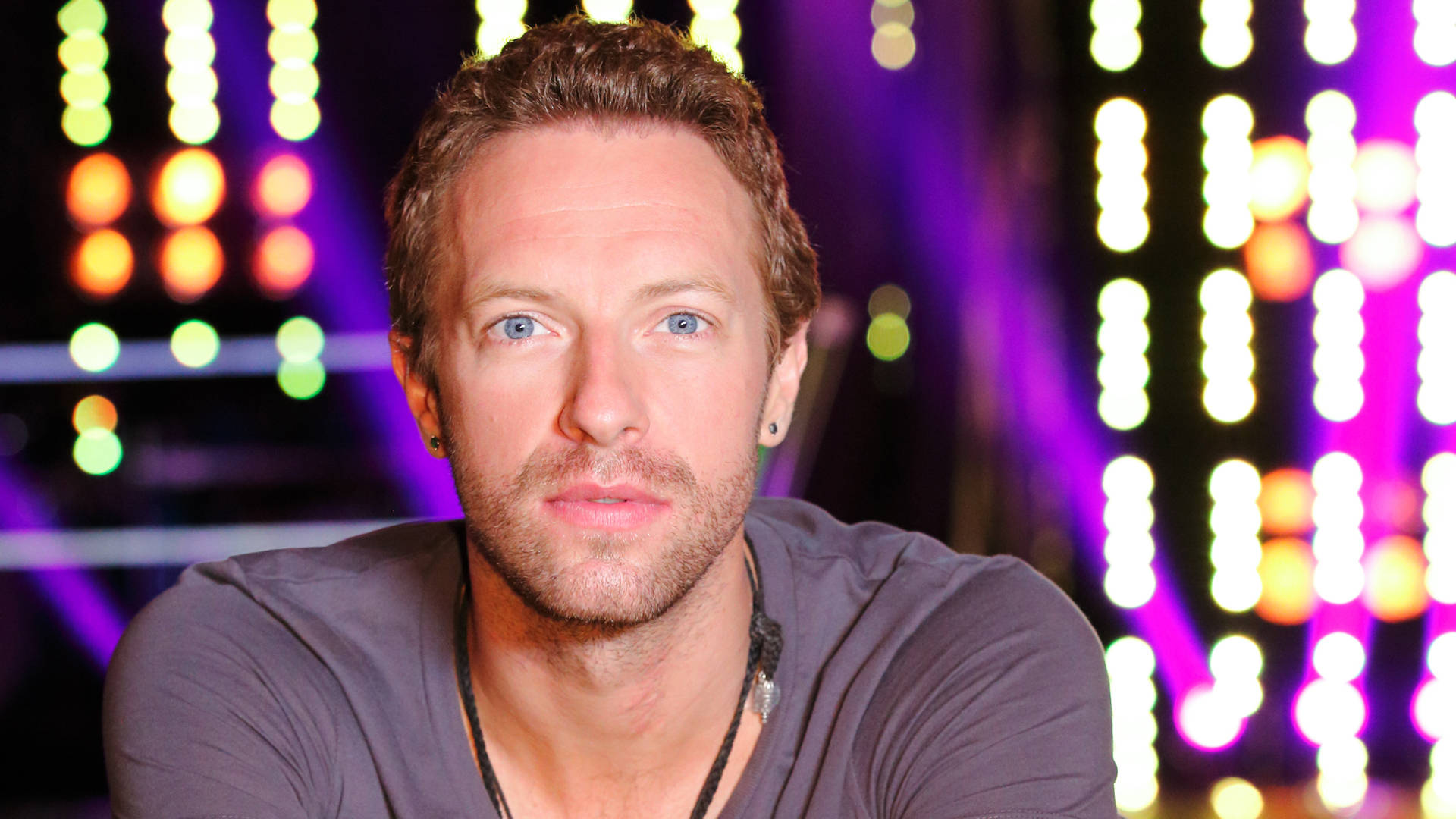 Chris Martin 桌布 壁紙 — 一個藍眼睛、短棕髮的男人直視鏡頭，背景是色彩繽紛舞台燈光形成的充滿活力的散景。
