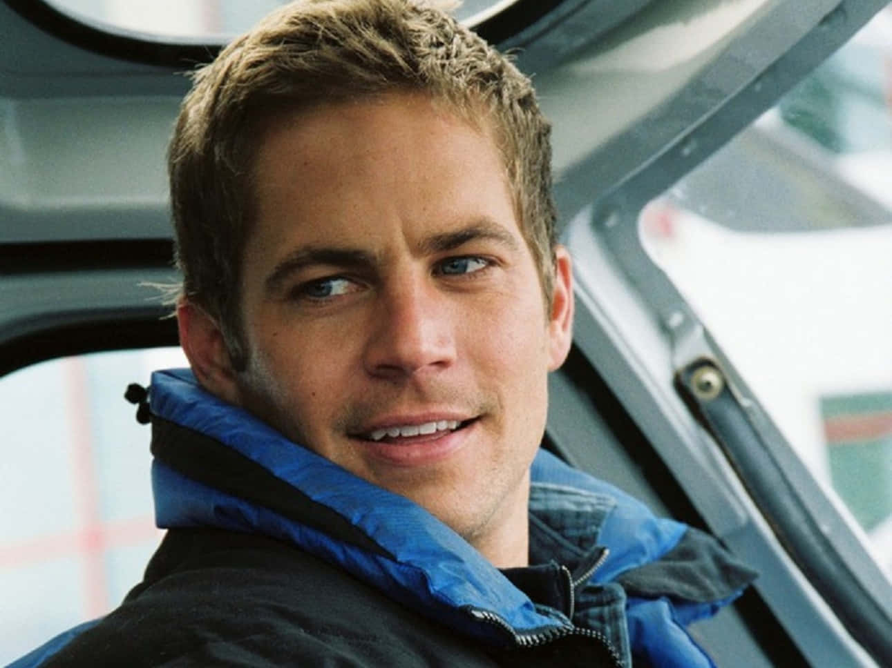 Paul Walker 桌布 壁紙 — 一個藍眼睛、短棕髮的男人穿著藍黑夾克，從車窗探出頭微笑。