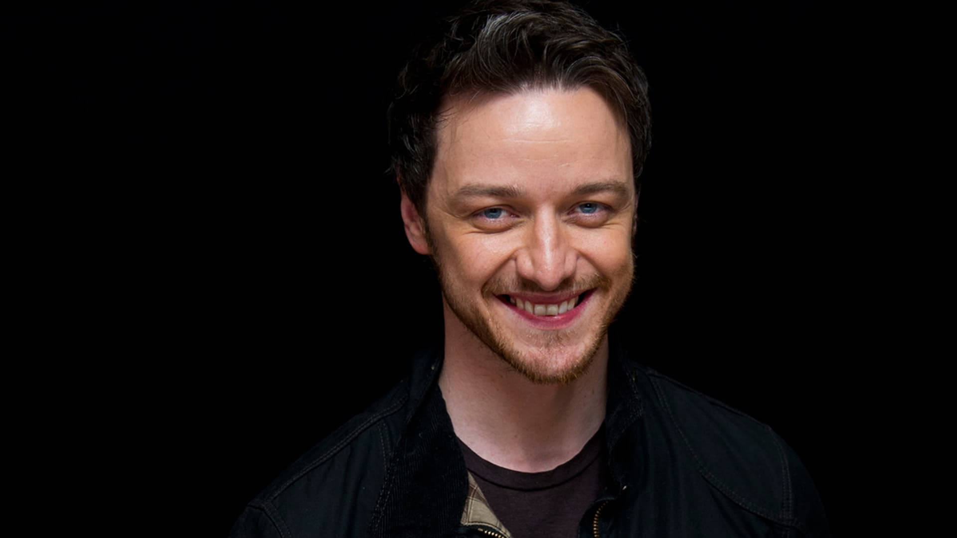 Celebrity 桌布 壁紙 — 一個藍眼睛、笑容溫暖的男人，穿著黑色外套，背景是純黑色。