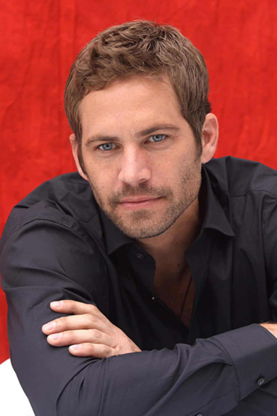 Paul Walker 桌布 壁紙 — 一個藍眼睛、鬍渣的男人，穿著黑色襯衫，在帶有紋理的紅色背景前身體前傾。
