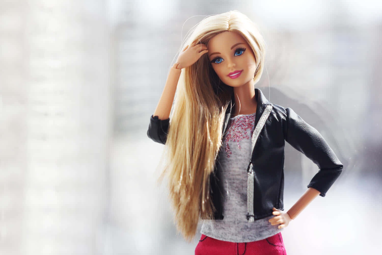 Barbie Doll 桌布 壁紙 — 一個藍眼睛的金髮娃娃穿著黑色外套，內搭灰色上衣和紅色褲子，一手叉腰，另一手觸摸頭髮擺姿勢。