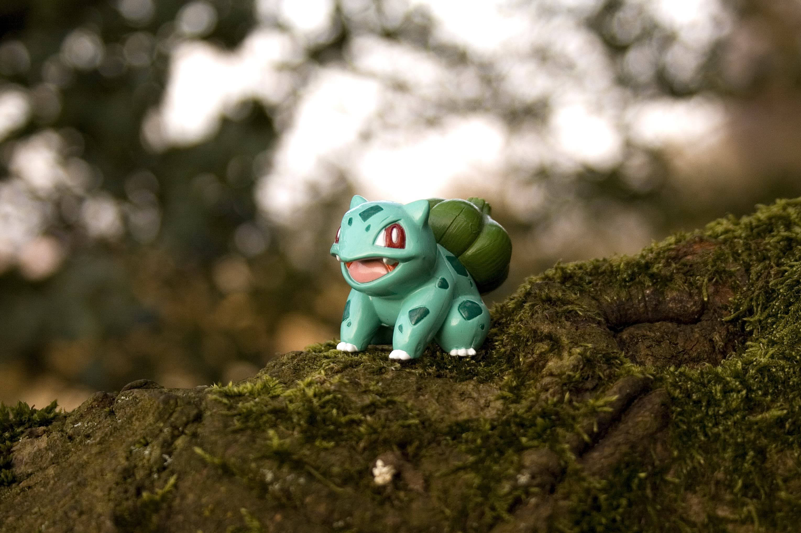 Bulbasaur 桌布 壁紙 — 一個藍綠色、紅眼睛的妙蛙種子玩具坐在長滿苔蘚的樹枝上，背景是模糊的森林。
