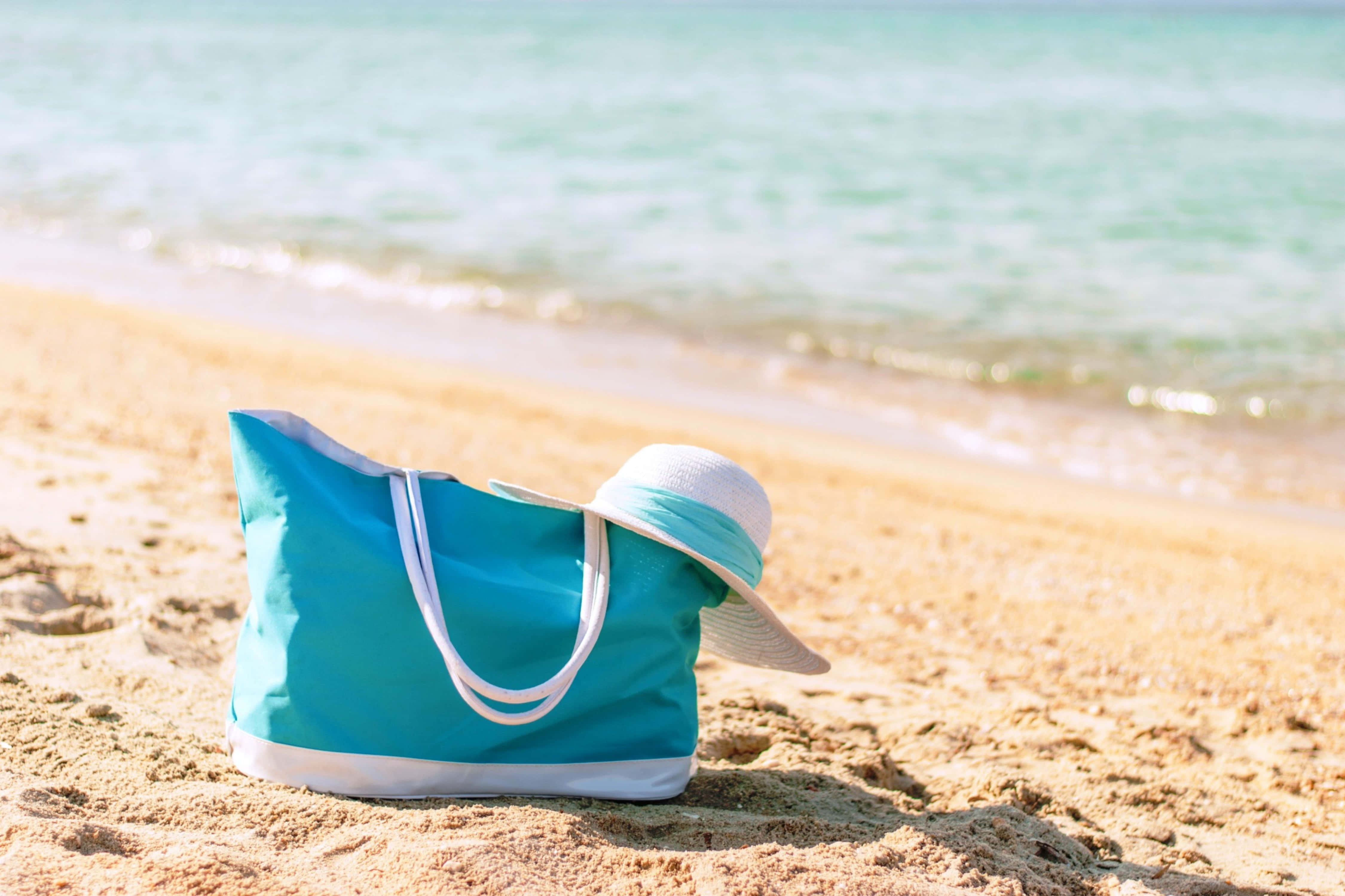 Beach Bag 桌布 壁紙 — 一個藍綠色沙灘袋和白色遮陽帽放在金色沙灘上，旁邊是柔和藍綠色波浪，陽光燦爛。