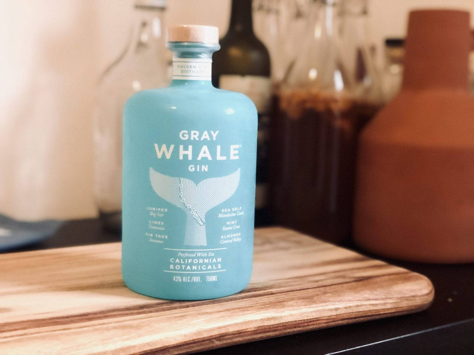 bottle 桌布 壁紙 — 一個藍綠色的Gray Whale Gin酒瓶放在木製砧板中央，背景有模糊的廚房用具。