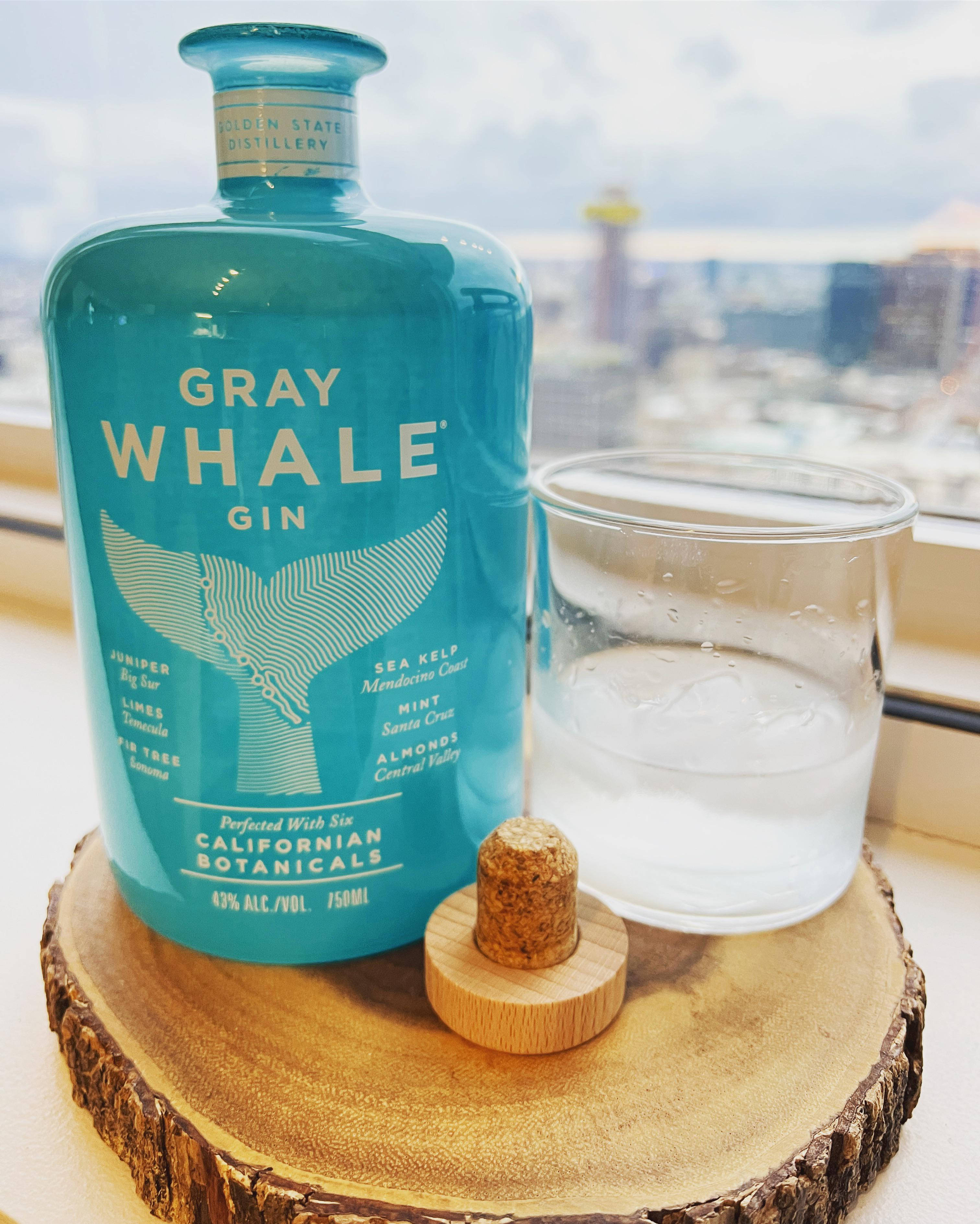 bottle 桌布 壁紙 — 一個藍綠色的Gray Whale Gin酒瓶放在木切片上，旁邊有一杯冰水，軟木塞放在杯墊上，透過窗戶俯瞰城市景觀。
