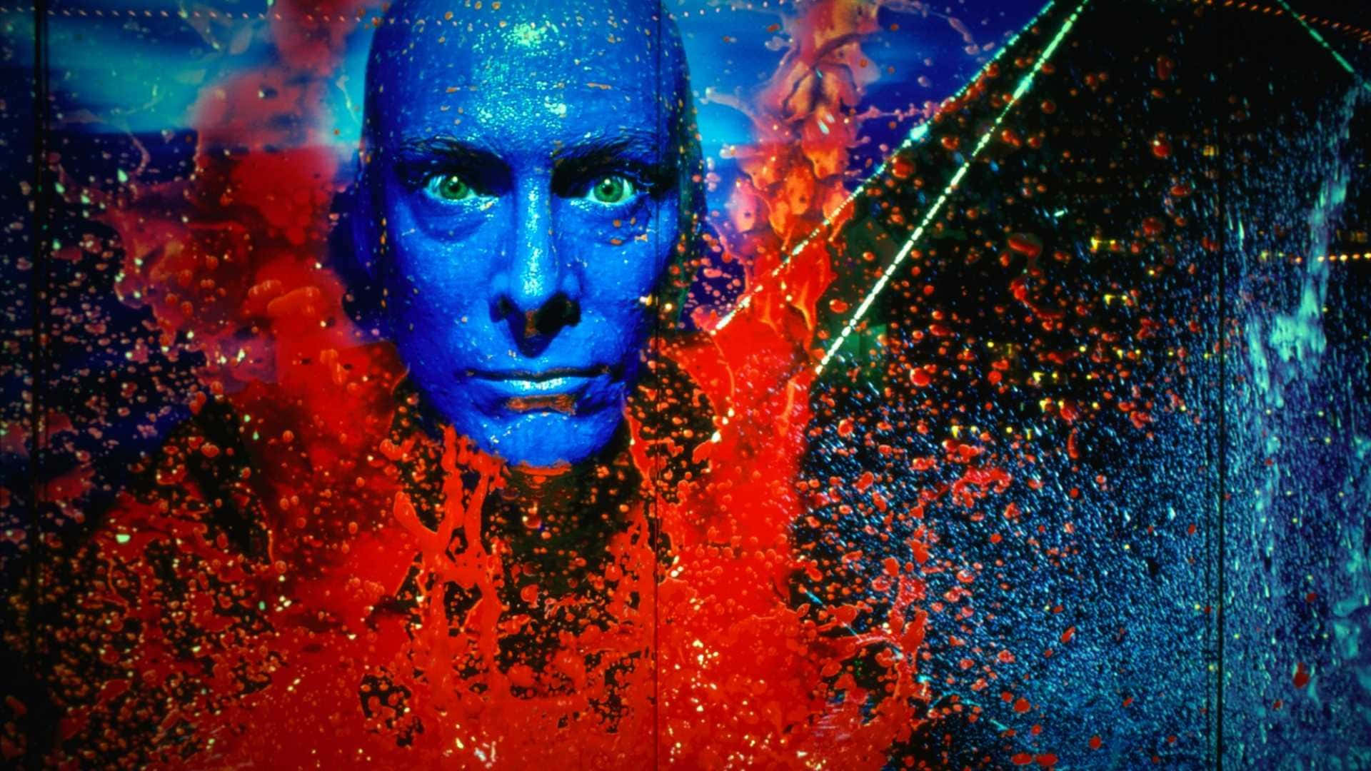 Blue Man Group 桌布 壁紙 — 一個藍臉人物，綠色眼睛，透過火紅和電藍色的抽象顏料和光爆炸凝視。