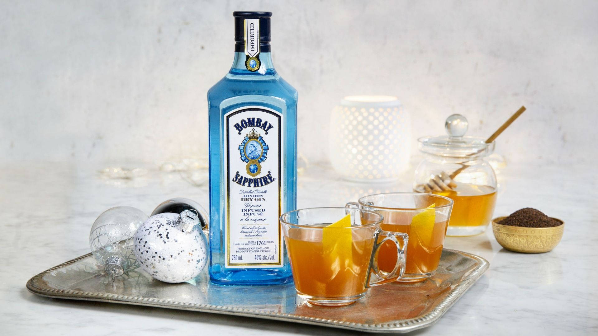 葡萄酒 桌布 壁紙 — 一個藍色Bombay Sapphire琴酒瓶放在銀托盤上，搭配兩杯蜂蜜柑橘飲品、節日裝飾品和一罐蜂蜜，背景是柔和的白色。