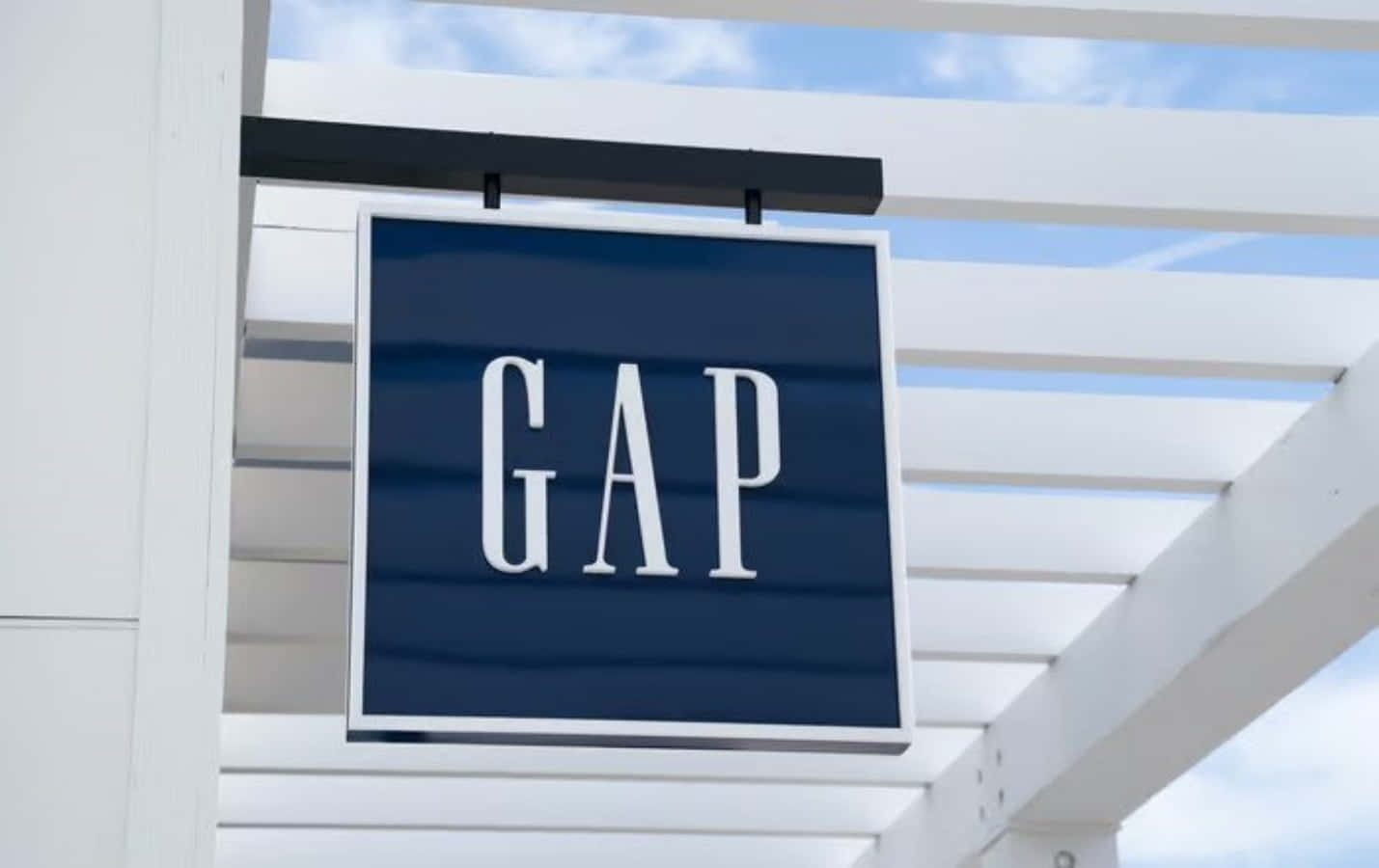 Gap 桌布 壁紙 — 一個藍色GAP商店招牌，白色字體，掛在白色涼棚下，背景是晴朗的藍天和零星白雲。